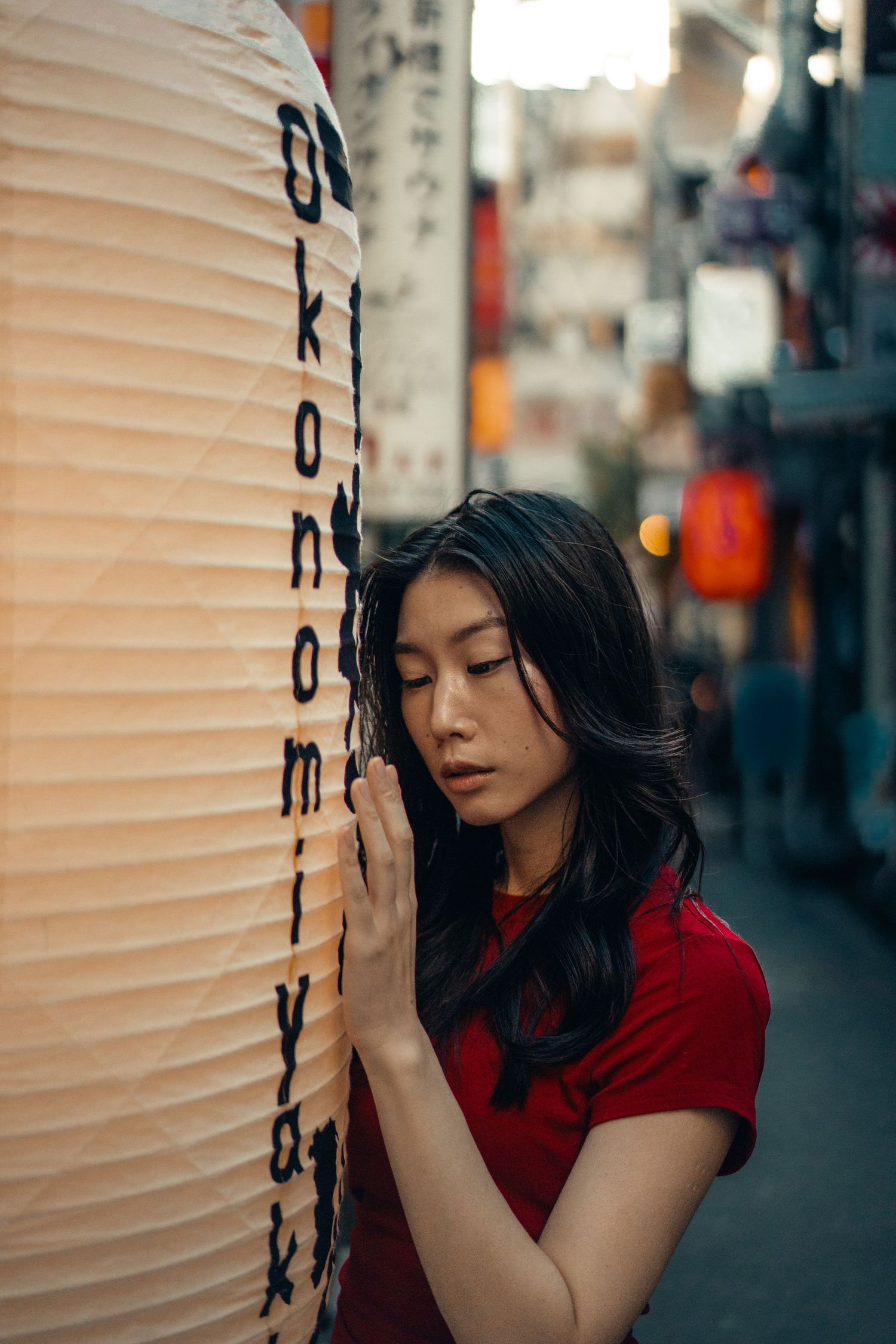 Woman in red shirt poses thoughtfully against white paper lanterns in moody urban street setting.