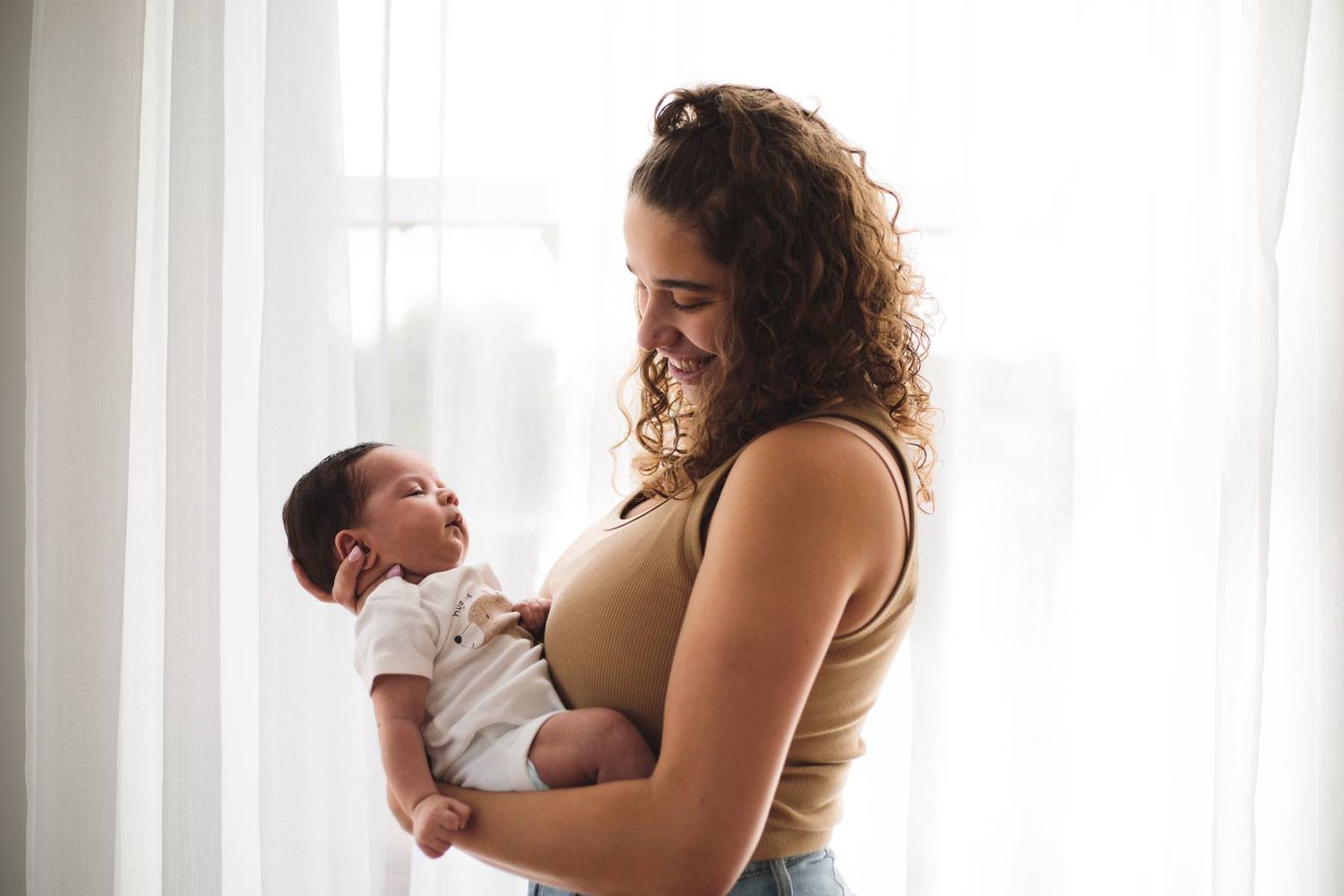 Smiling mother with curly hair gazes lovingly at her newborn baby held in her arms beside a sunlit window.