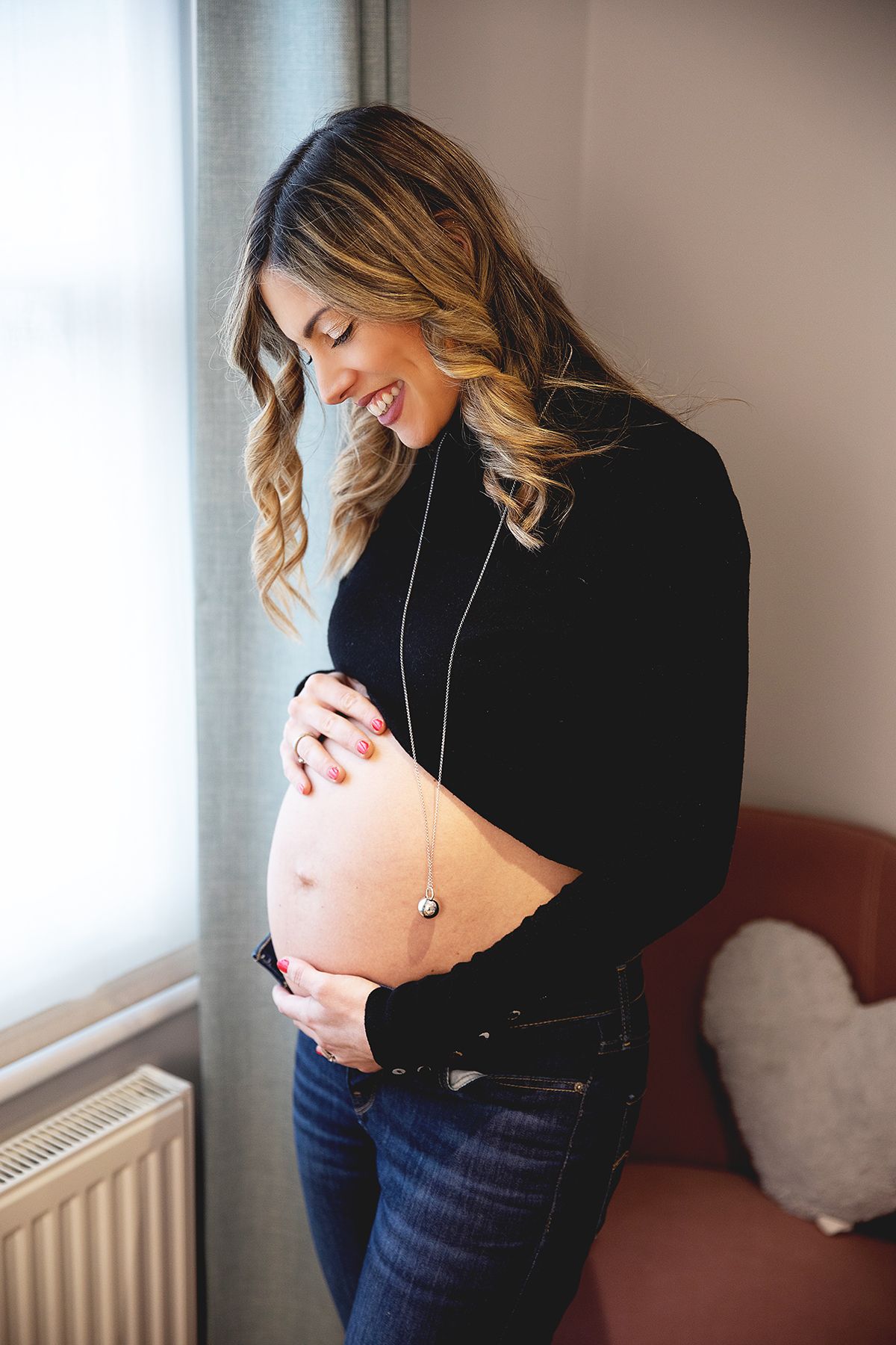 Expectant woman in black crop top and jeans smiles while touching her pregnant belly near a window. During a photoshoot in London