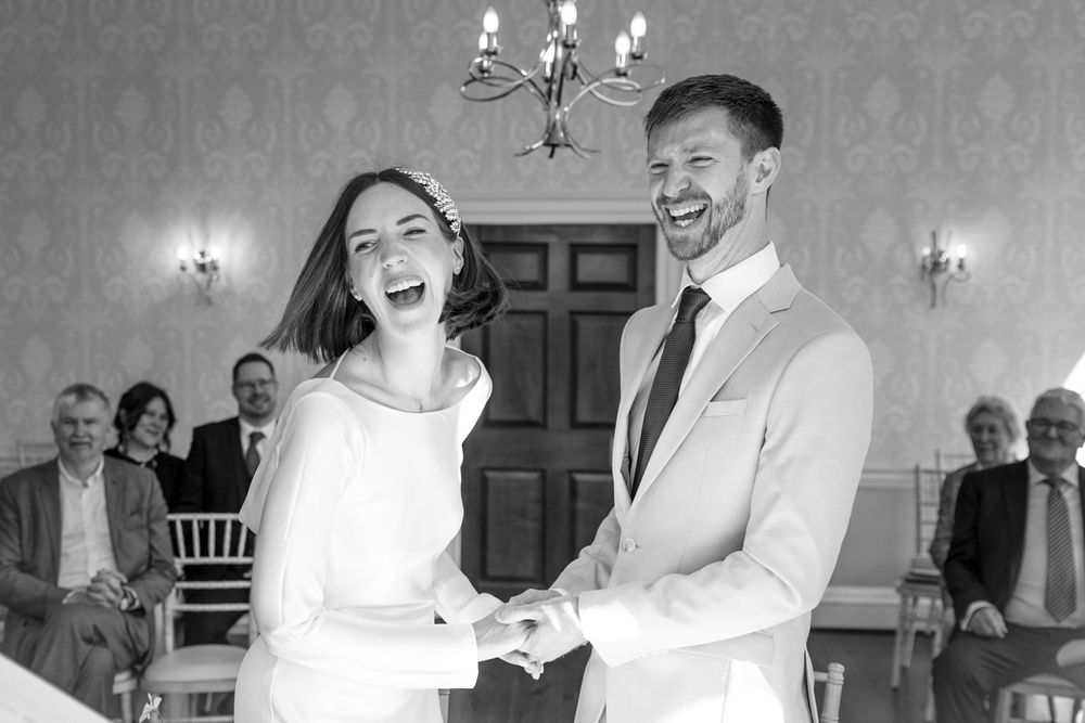 A joyful couple laughs together during their wedding ceremony while guests look on in the background.
