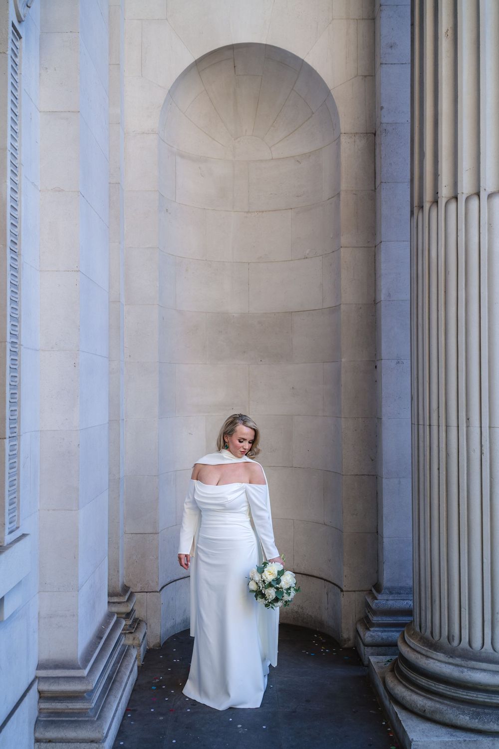 Bride in elegant white off-shoulder gown holding bouquet, posed between classical stone columns and arched alcove.
