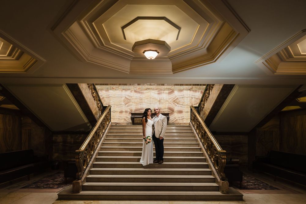 Couple embraces on grand staircase in elegant historic building with ornate ceiling and warm lighting.