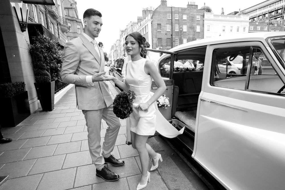 Black and white photo of a fashionable vintage couple stepping out of a classic car in an urban setting.