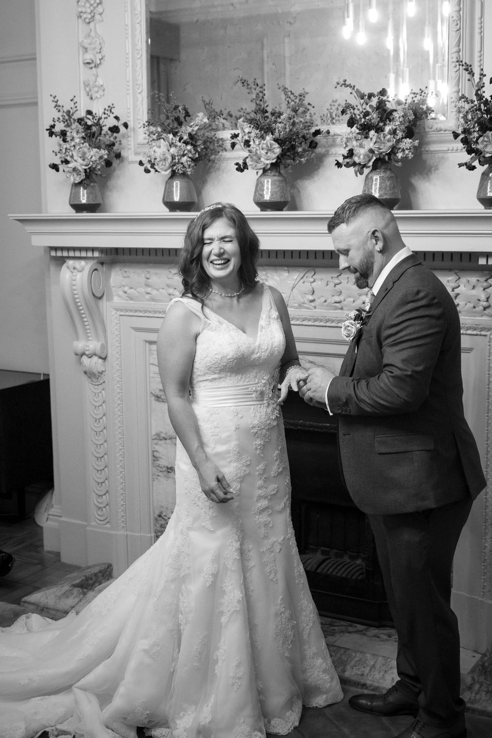 Black and white photo of a laughing bride in a lace gown and groom in a suit exchanging rings by an elegant fireplace.