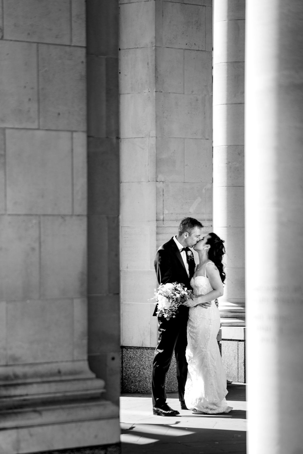 A romantic black and white wedding photo of a couple embracing between elegant stone columns.