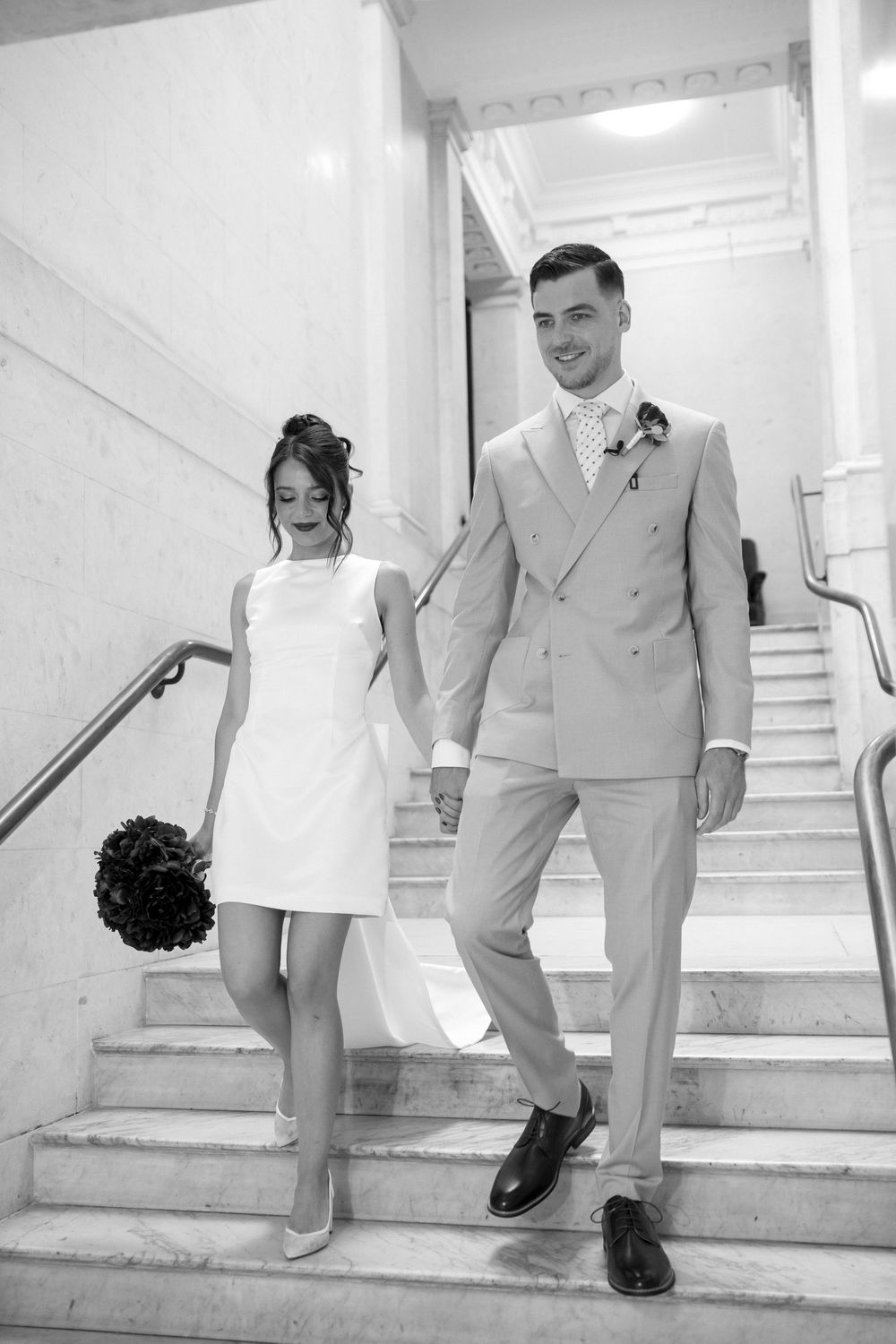 A bride in a short white dress and groom in a light suit descend marble stairs while holding hands in black and white.