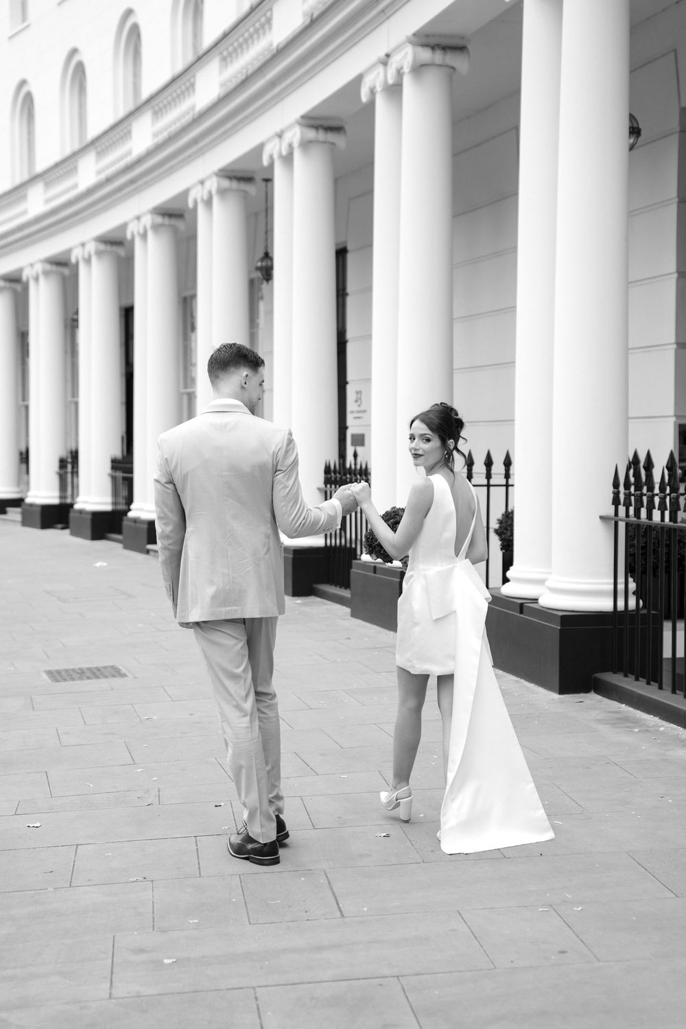 A wedding couple stands between white architectural columns during a photo session on a stone walkway.