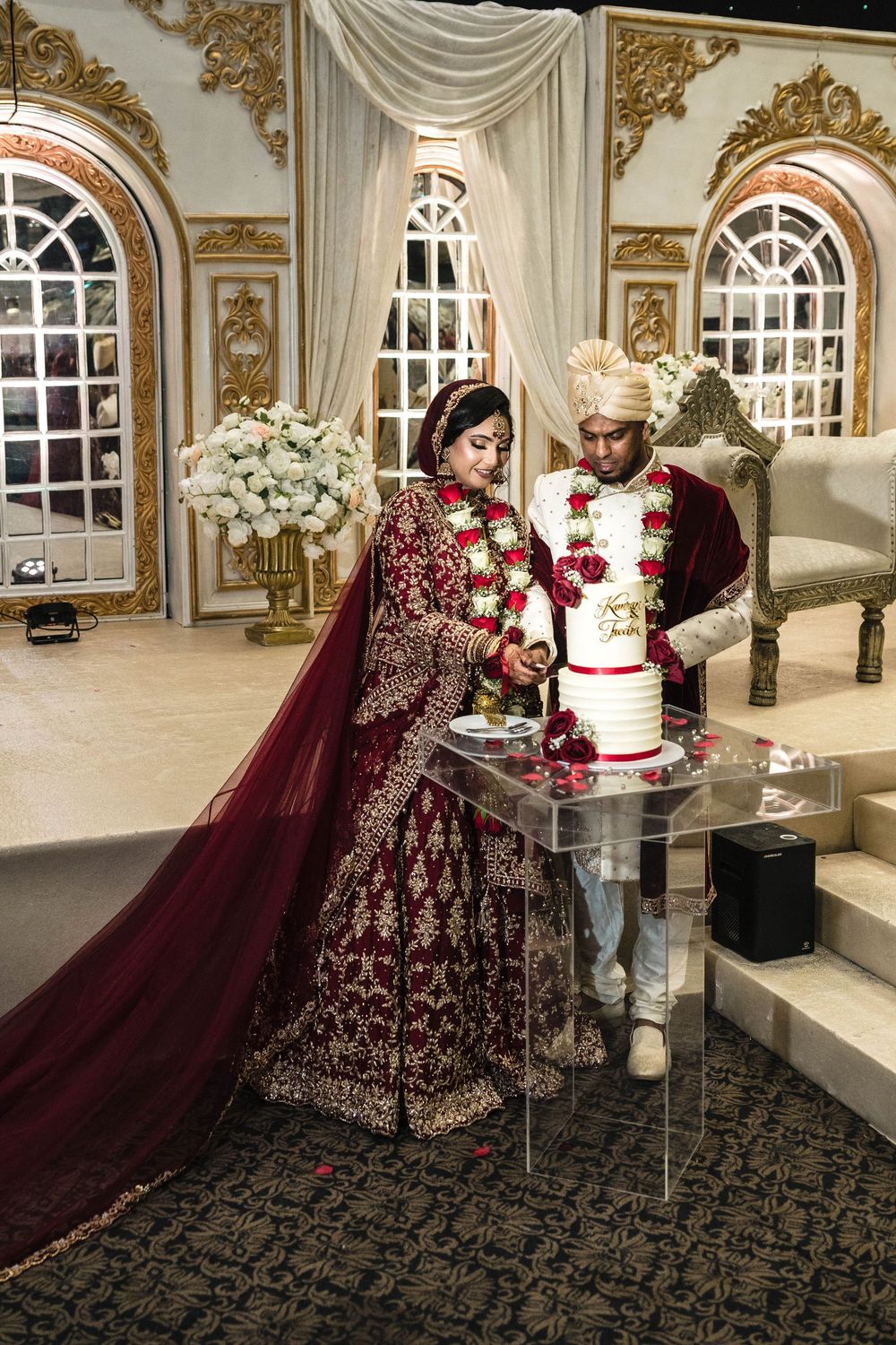 Indian bride and groom in traditional red attire cutting wedding cake at elegant gold-decorated venue.