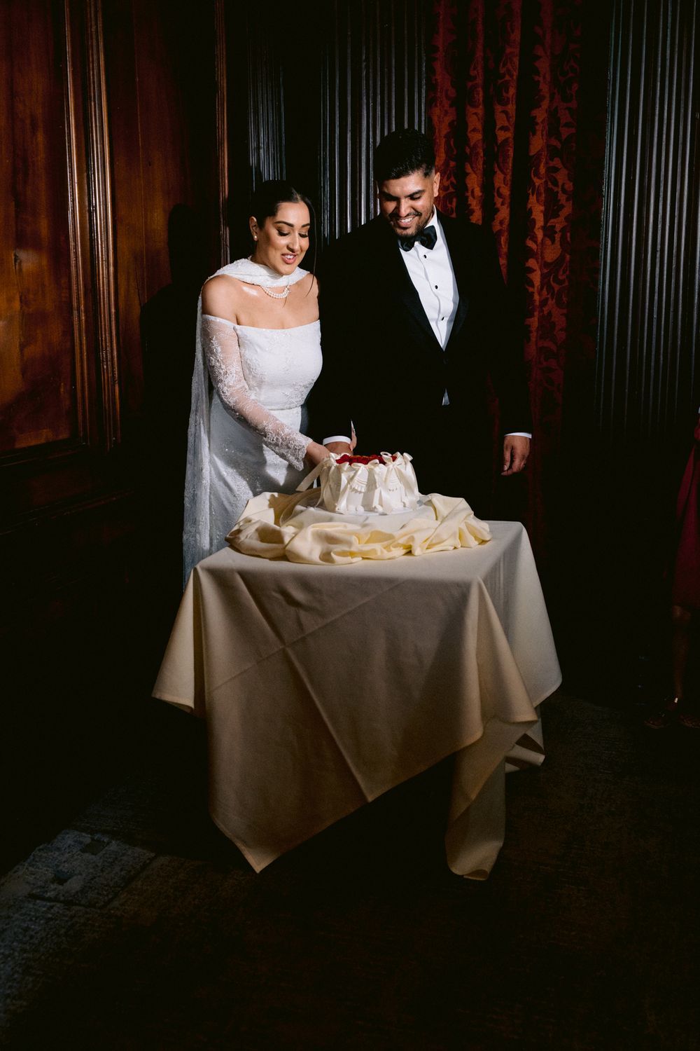 A couple in formal wedding attire stands at an elegantly decorated table with a wedding cake at a dark wood-paneled venue.