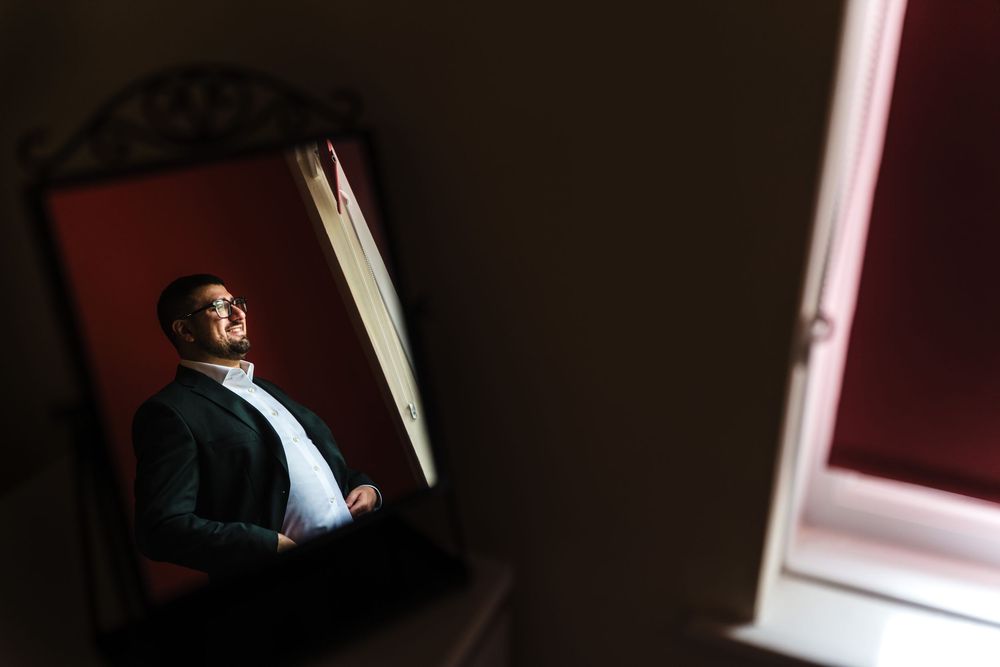 A person in formal attire stands by a window, illuminated by dramatic lighting against a dark red wall.