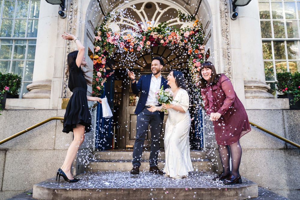 Shower of confetti falls on newlyweds and guests on ornate stone steps of historic building entrance.