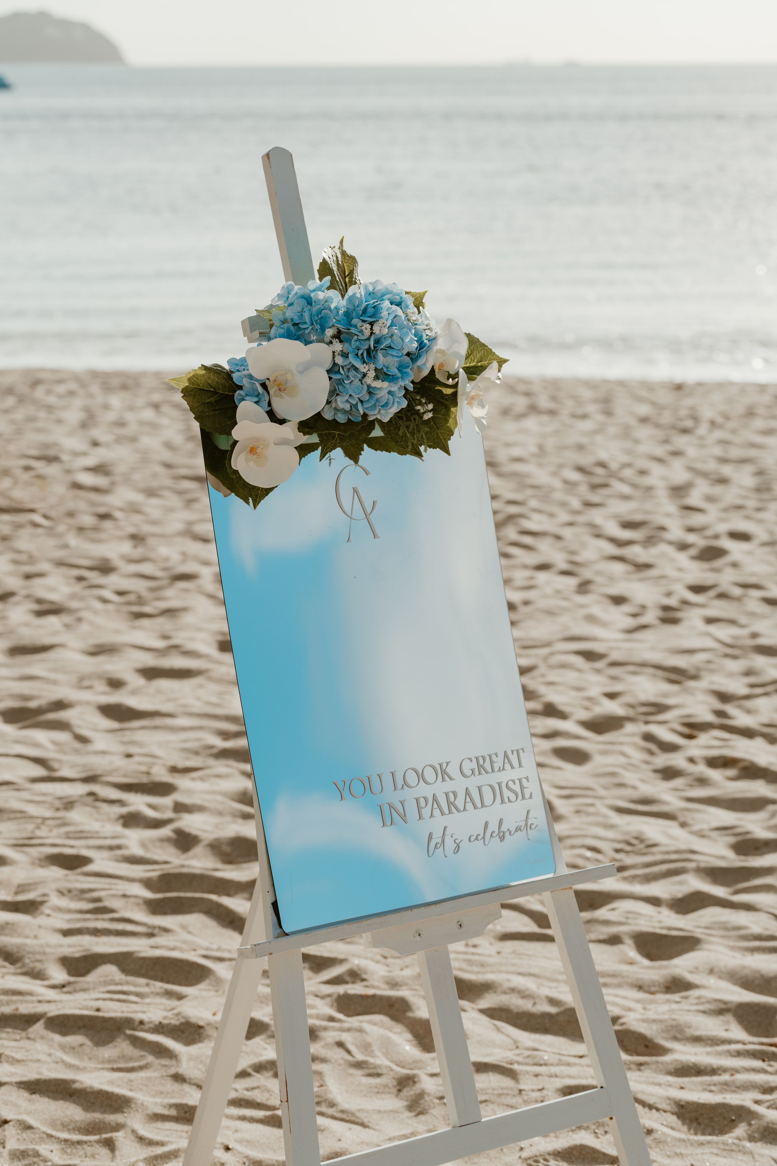 Beach wedding welcome sign on easel with blue hydrangeas reading You Look Great in Paradise.