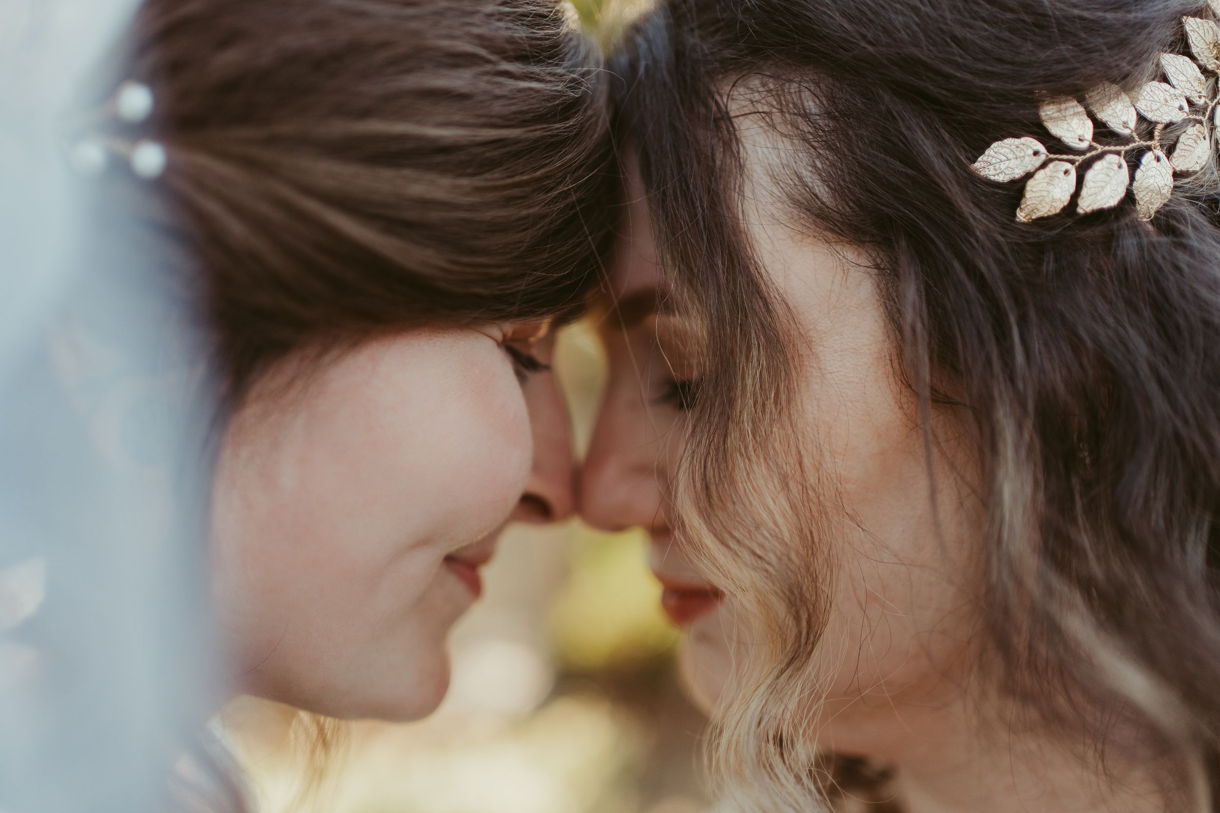 Two brides share an intimate moment, touching foreheads, one wearing a gold leaf hair accessory on their wedding day.