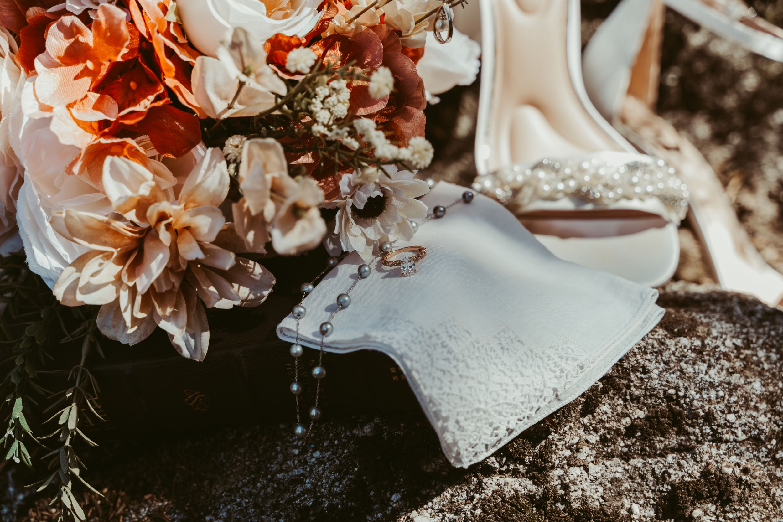 Bridal details flat lay with orange bouquet, wedding rings, rosary, and embellished heels on rock.