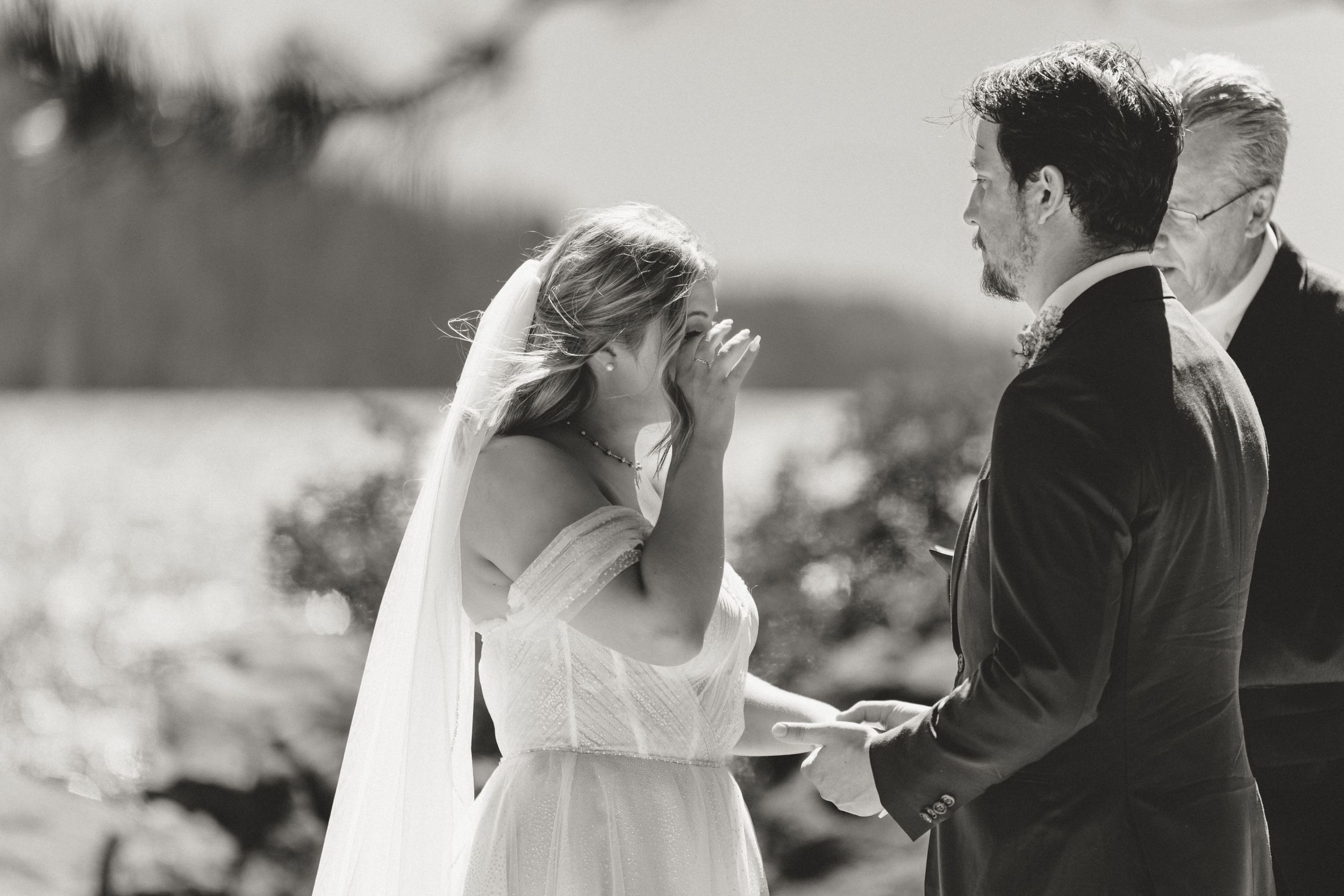 Emotional black and white photo of bride wiping tears during outdoor lakeside wedding ceremony.