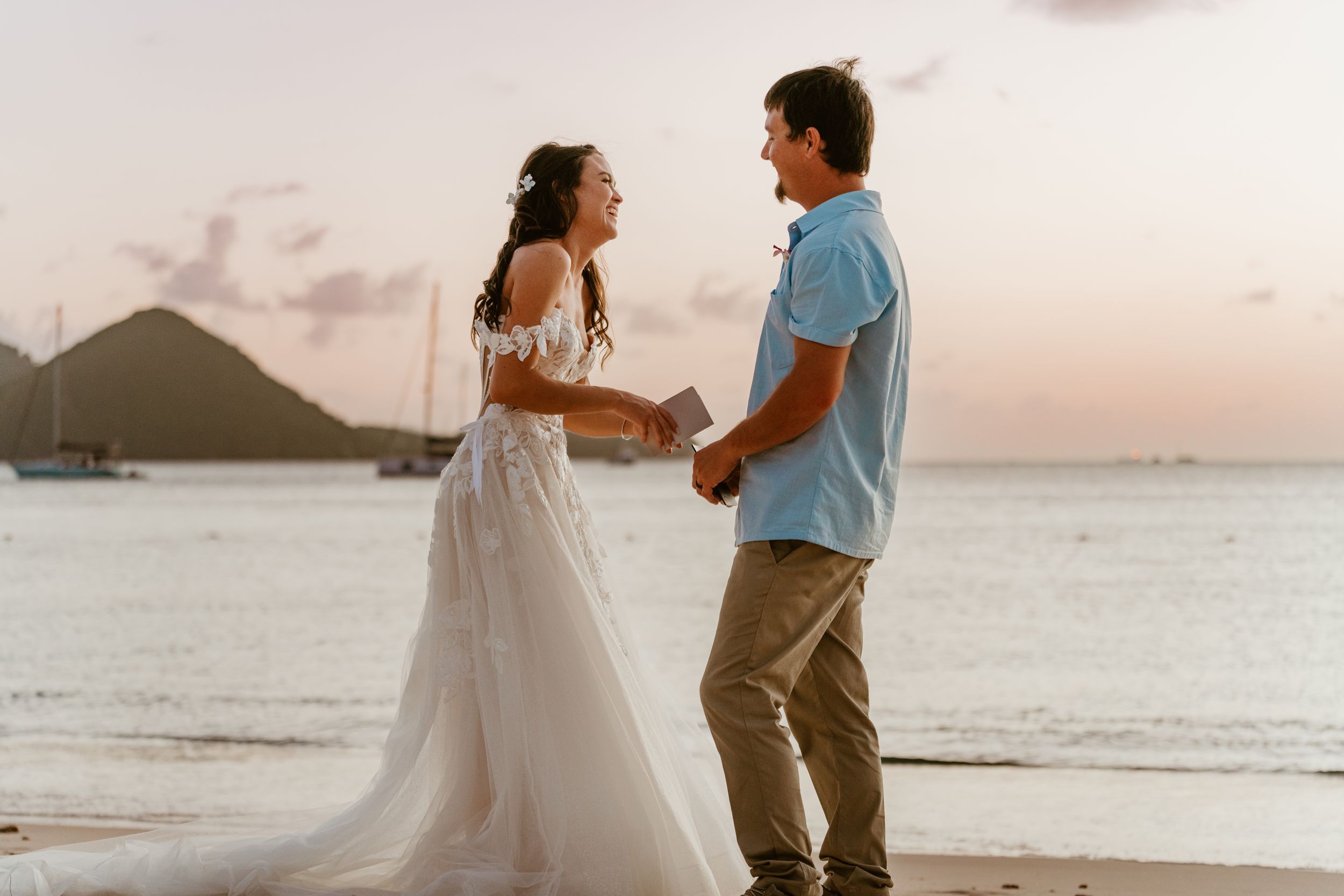 Bride and groom holding hands on a tropical beach at sunset with sailboats in the background.