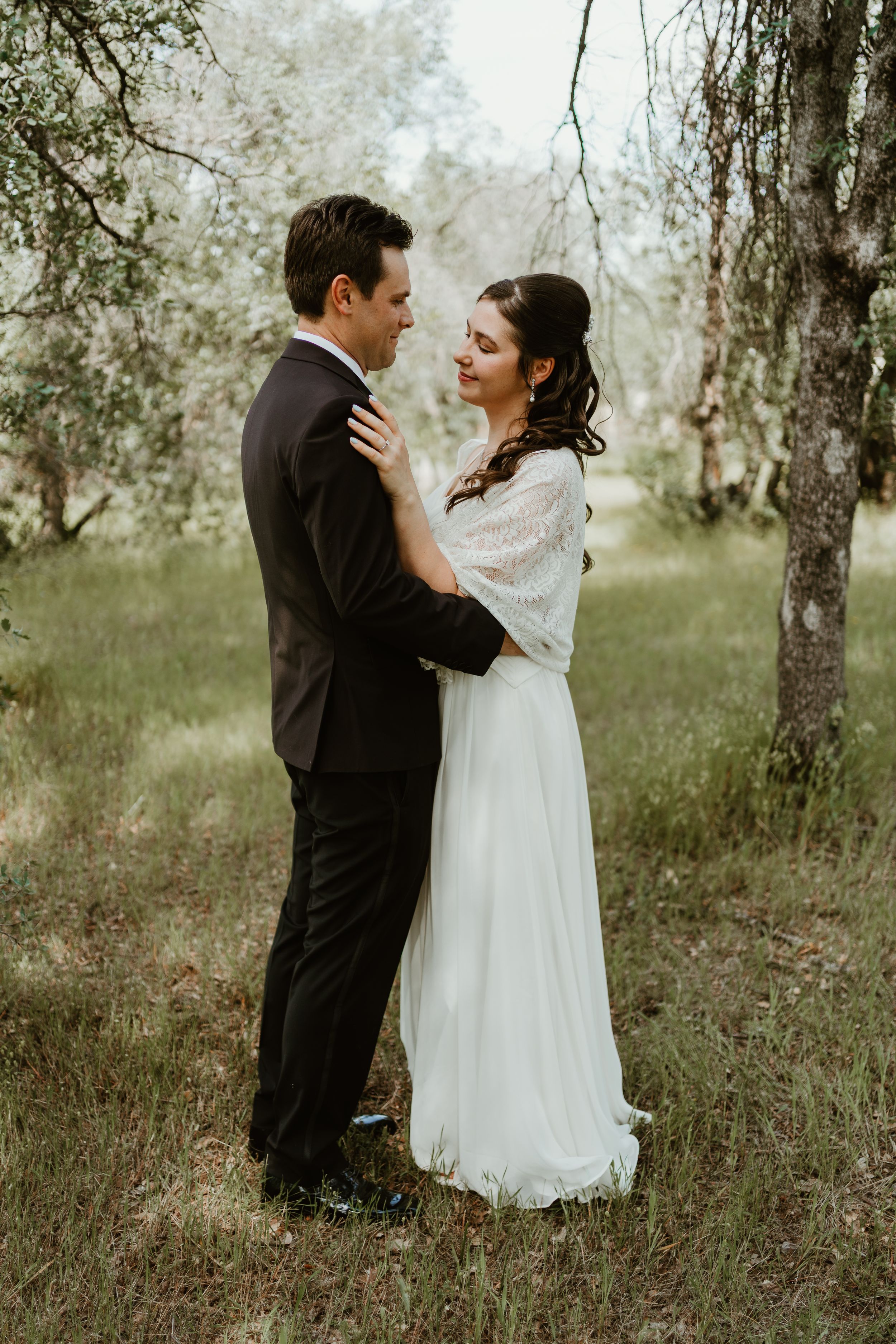 A bride and groom embrace romantically in a lush green outdoor setting, with the bride wearing a flowing white lace dress.