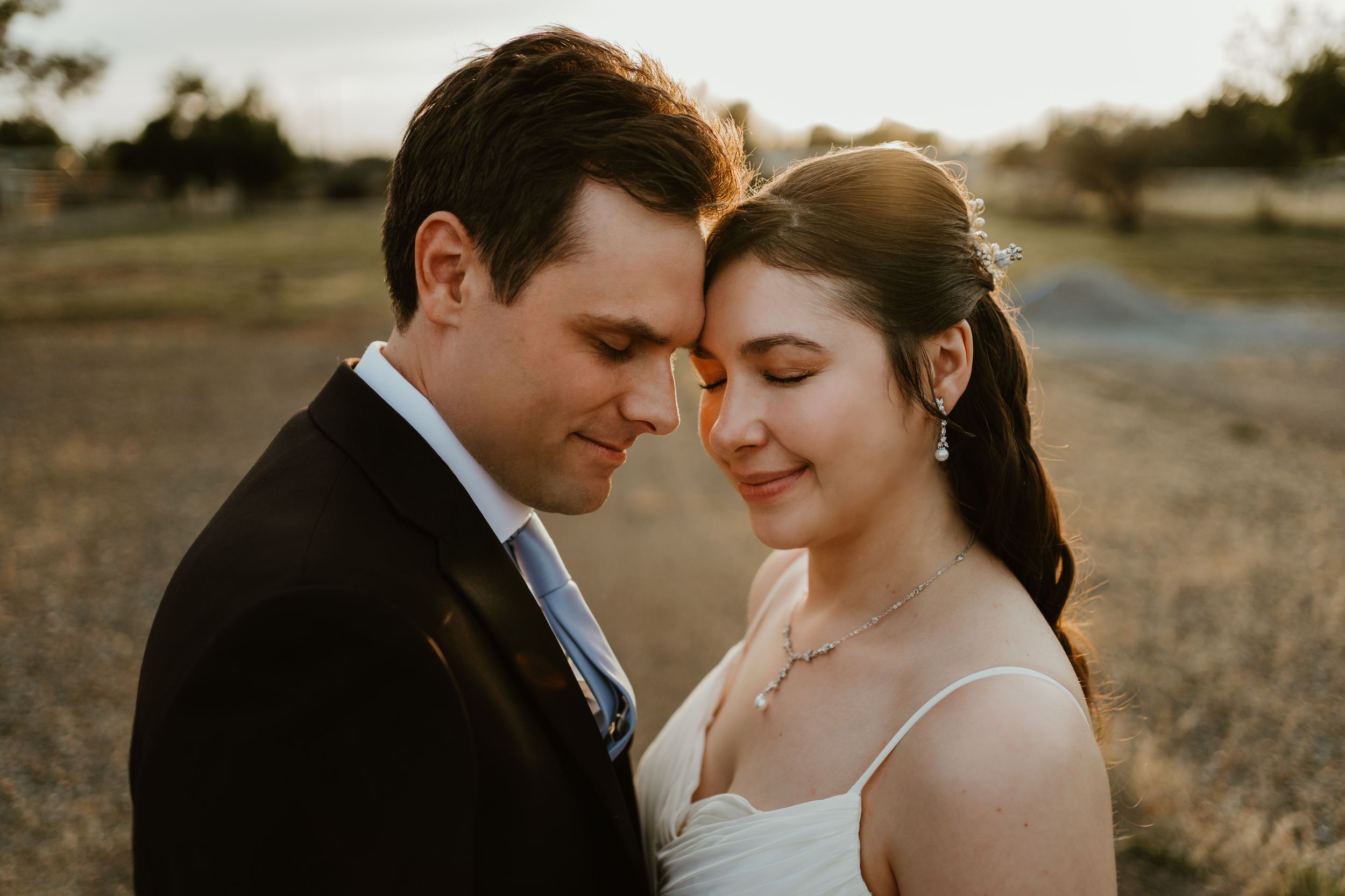 A bride and groom share an intimate forehead touch at golden hour in a rustic field setting.
