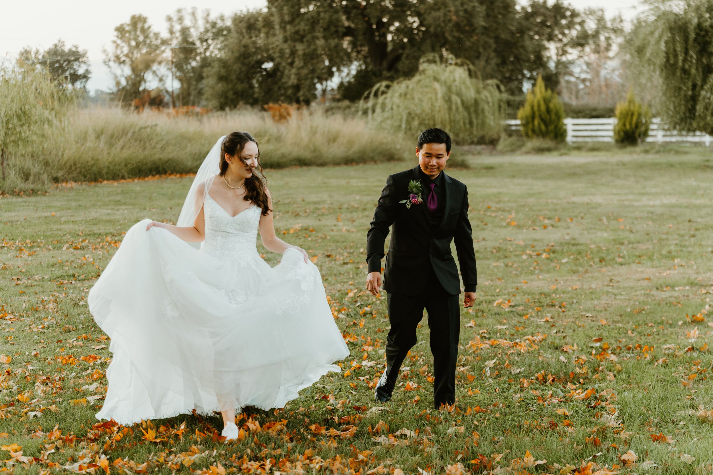 Bride in flowing white gown and groom in black suit walk hand in hand on autumn lawn with colorful fallen leaves.