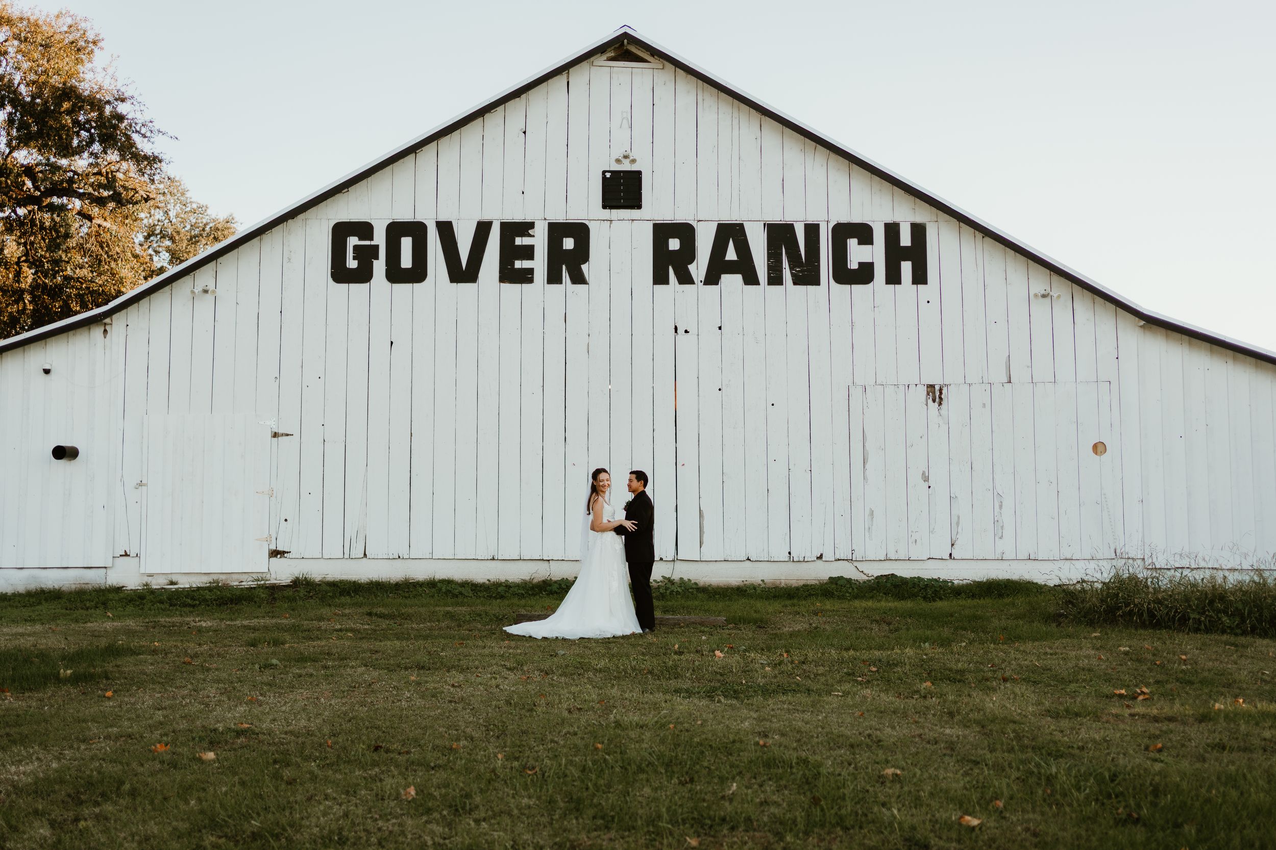 Bride and groom stand before a large white barn with Gover Ranch sign on a sunny day.