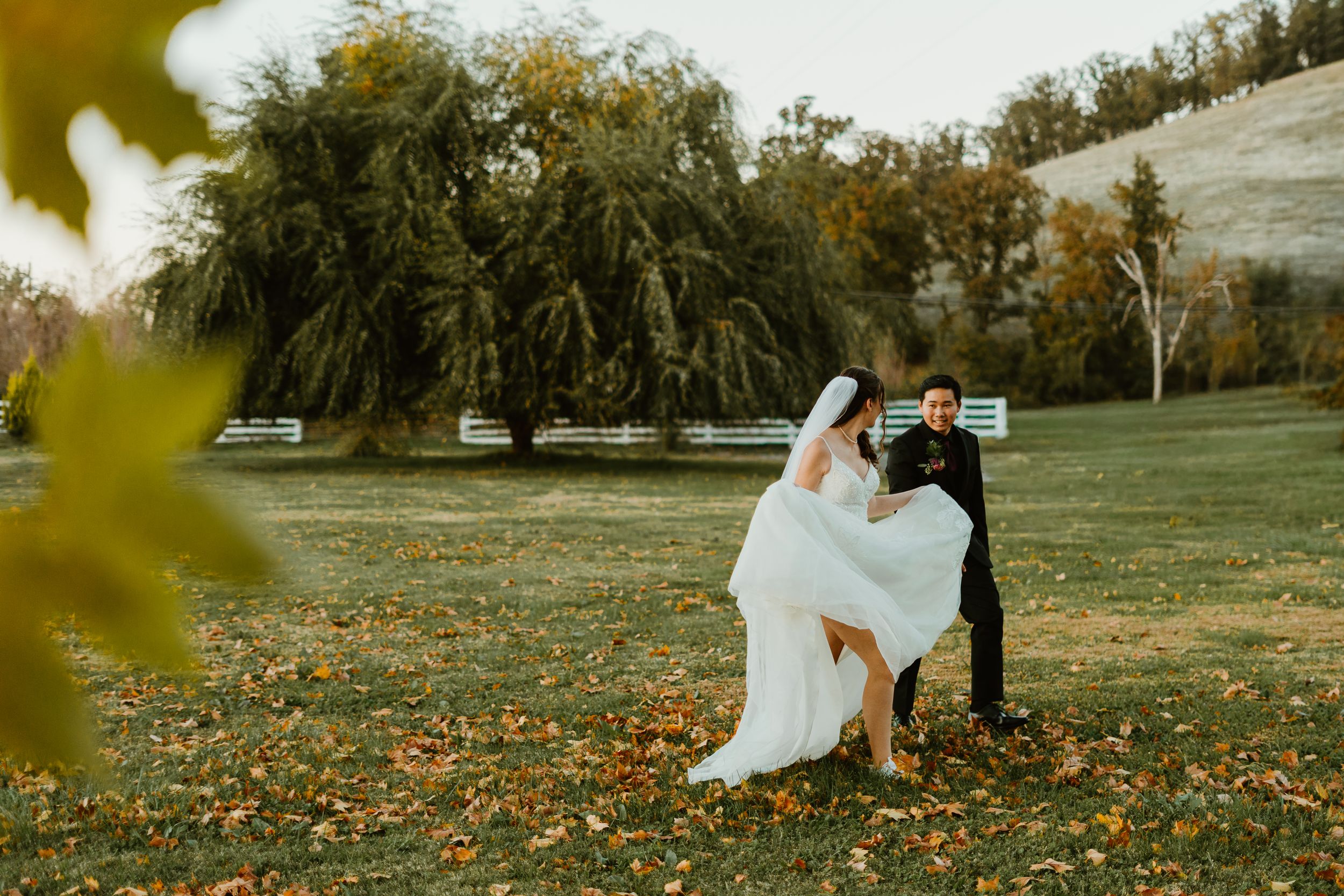 Bride and groom share a romantic moment on a park bench surrounded by autumn foliage and weeping willow trees.