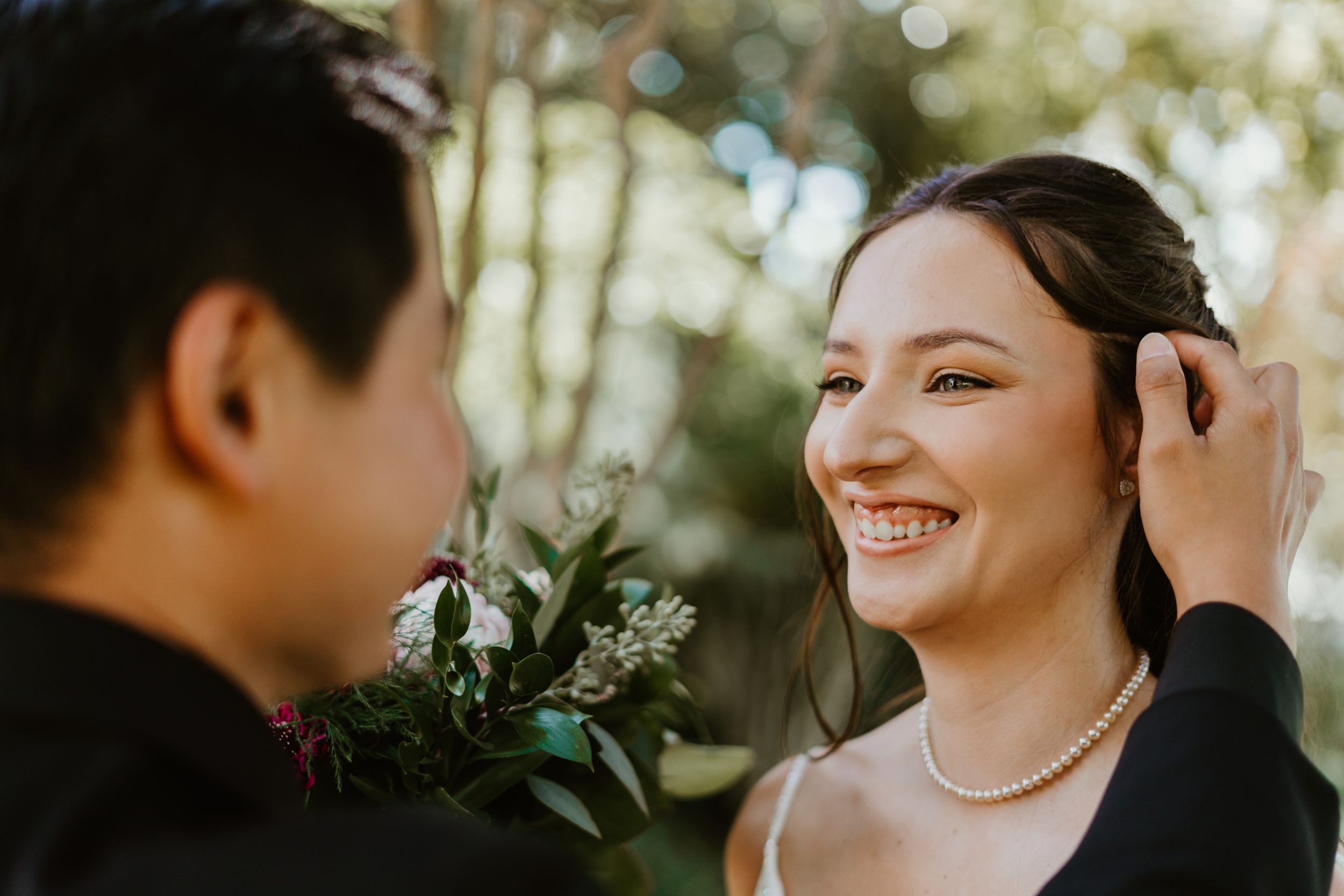 A smiling bride in pearl necklace gazes at groom holding floral boutonniere during outdoor wedding ceremony.