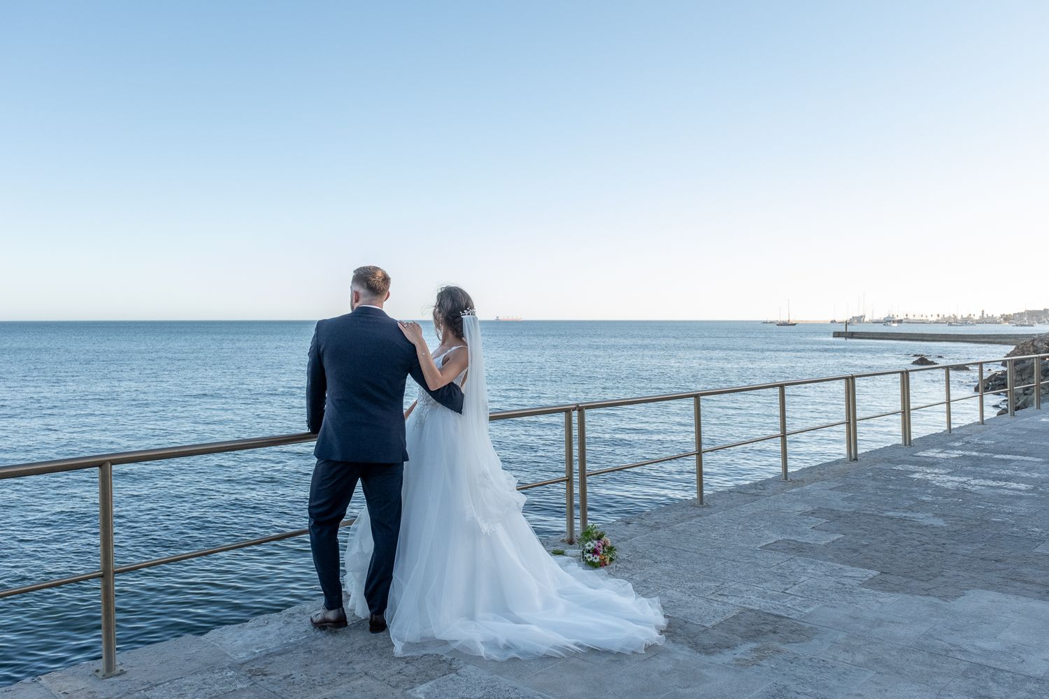 Wedding couple walking along oceanfront boardwalk with scenic water views.