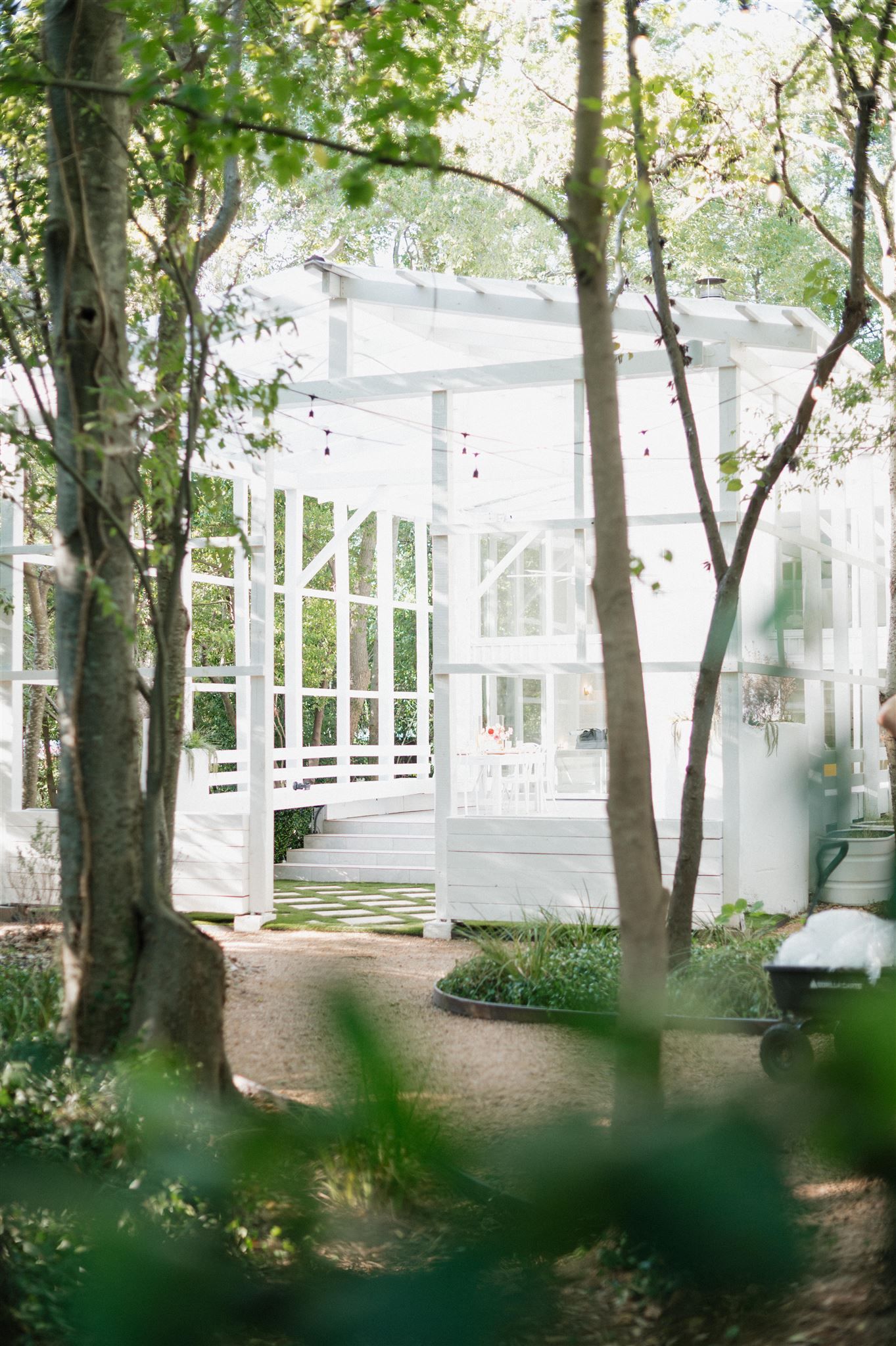 A white wooden gazebo or conservatory structure nestled among trees in a dreamy garden setting.
