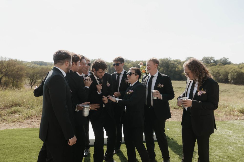 Groomsmen in black suits laughing and socializing outdoors before a wedding ceremony.