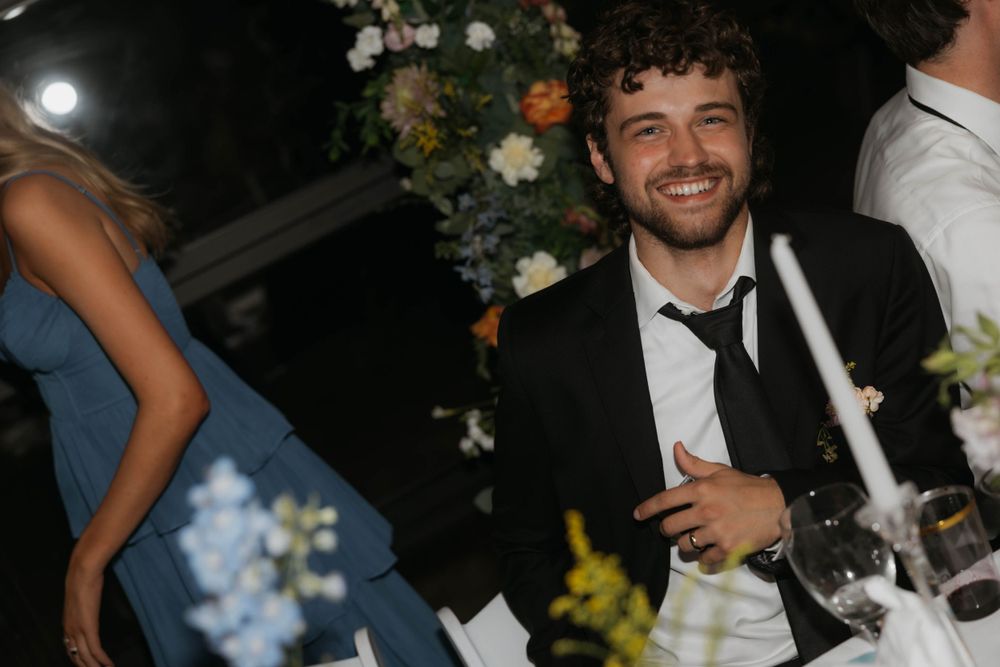 Happy groom in black suit smiling at decorated wedding reception dinner table.