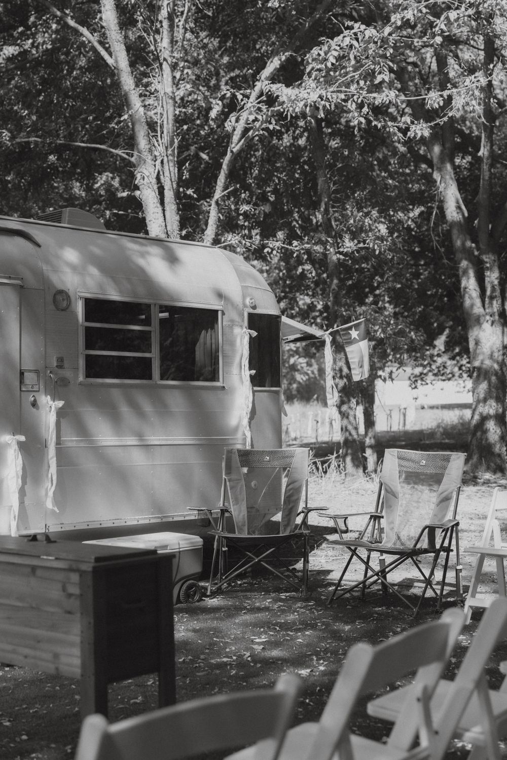 Black and white photo of vintage silver travel trailer with folding chairs at rustic outdoor wedding venue.