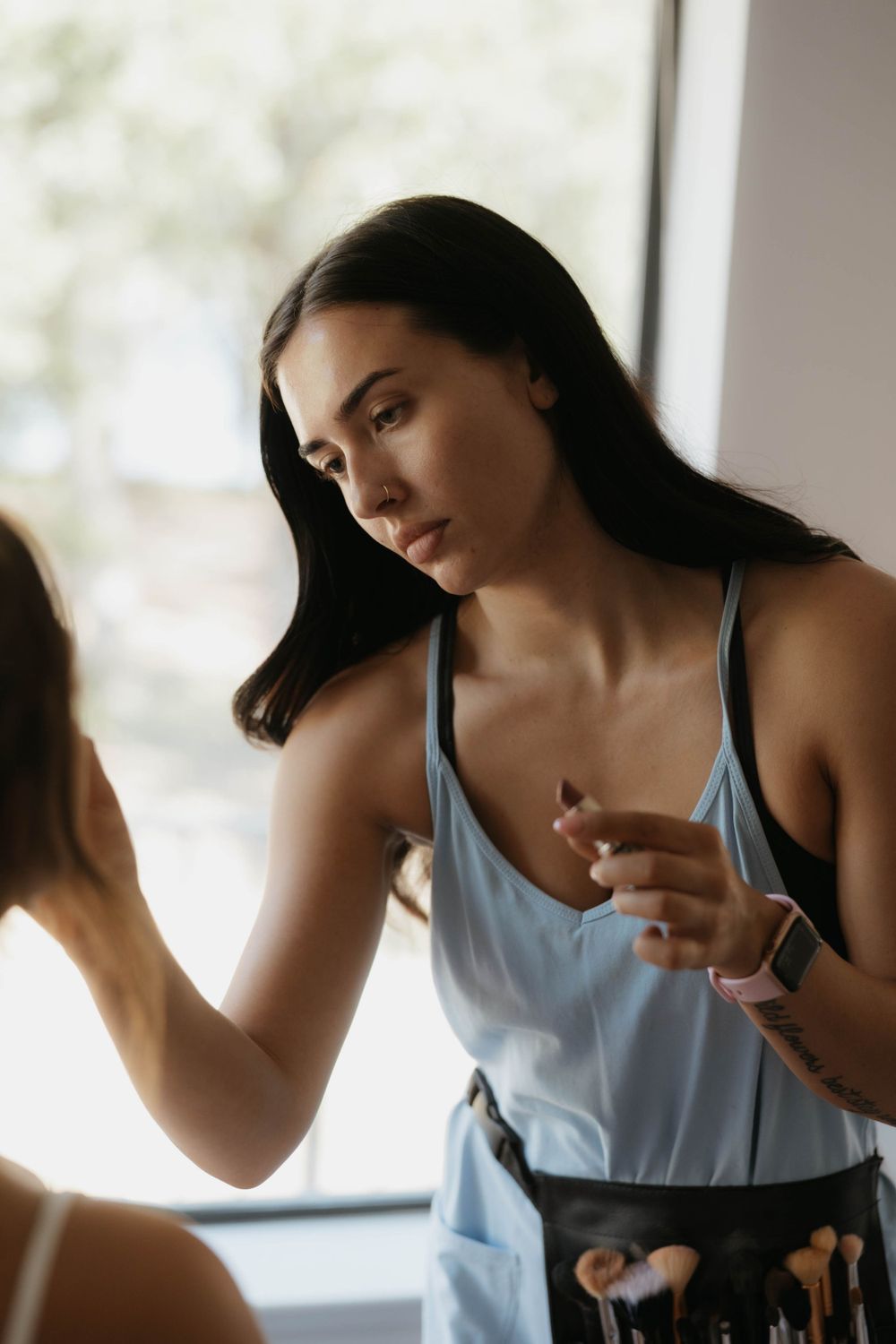 Professional makeup artist focusing while applying makeup to client near bright window.