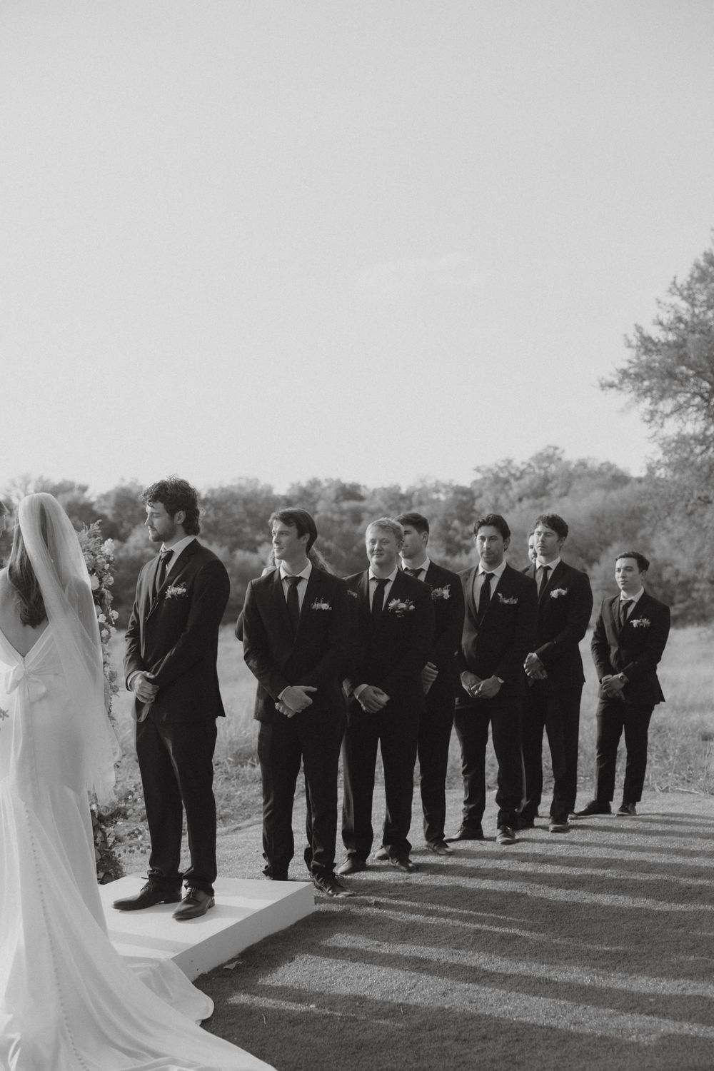 Black and white photo of groomsmen in dark suits standing at outdoor wedding ceremony altar.