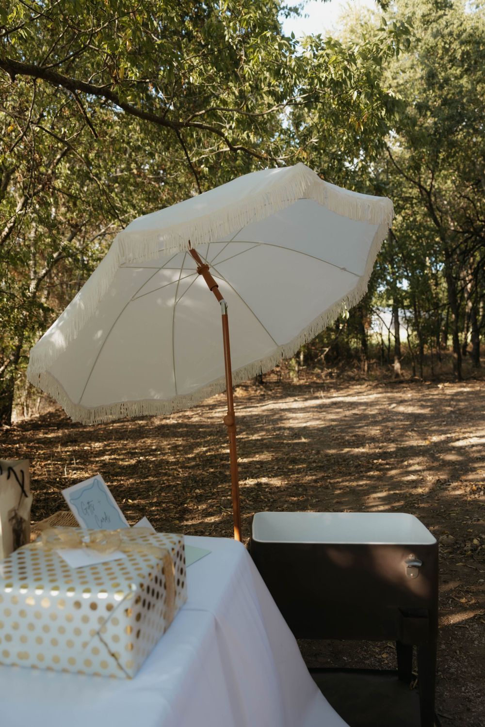 White fringed patio umbrella shading gift table with gold polka dot present at outdoor woodland wedding.