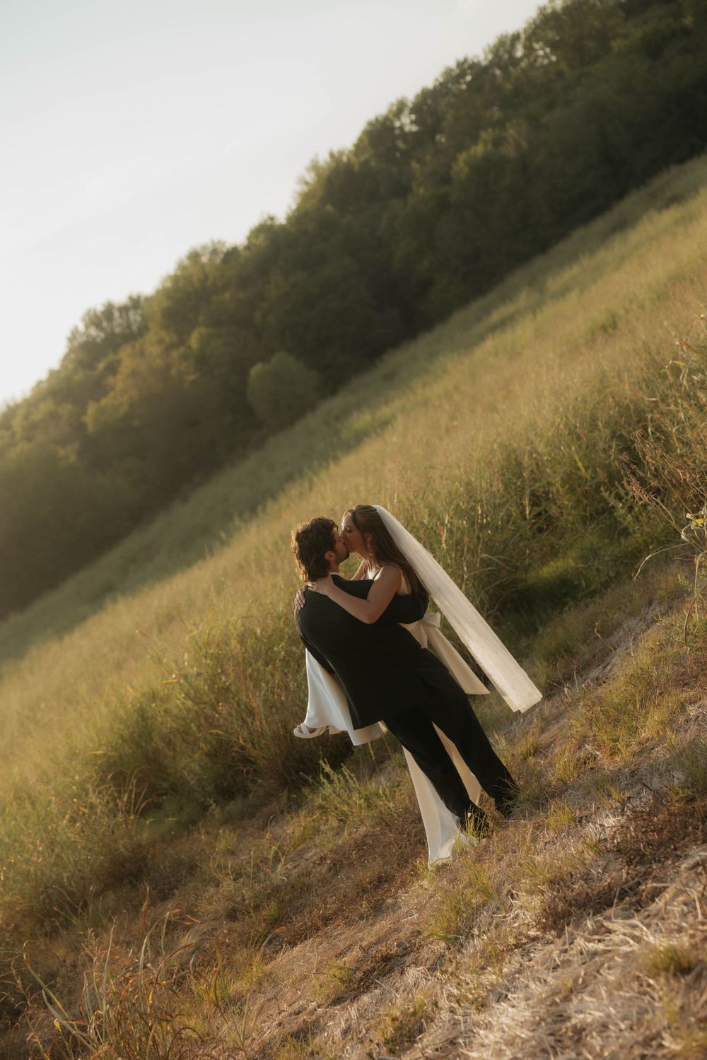 Bride and groom kissing in a sunlit green meadow field during golden hour wedding portrait session.