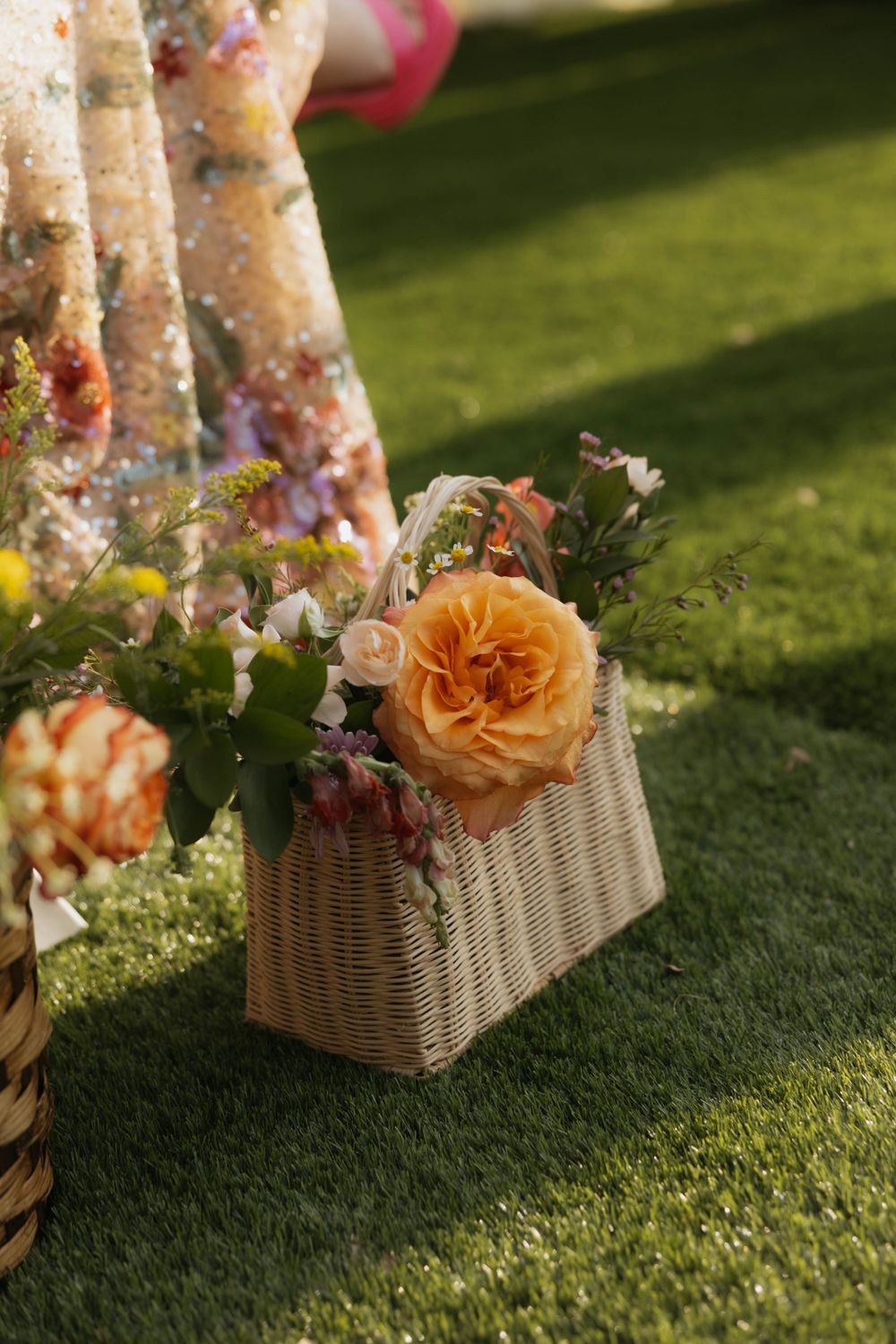 Wicker basket filled with colorful wildflowers and a peach garden rose sitting on green grass at an outdoor wedding.