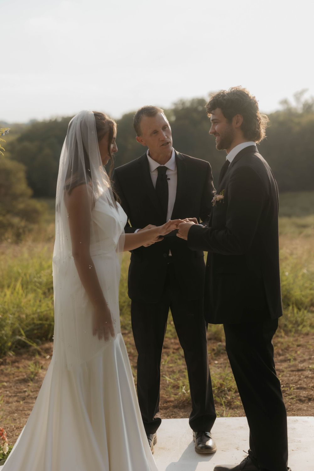Bride and groom exchanging rings during golden hour outdoor wedding ceremony with officiant.
