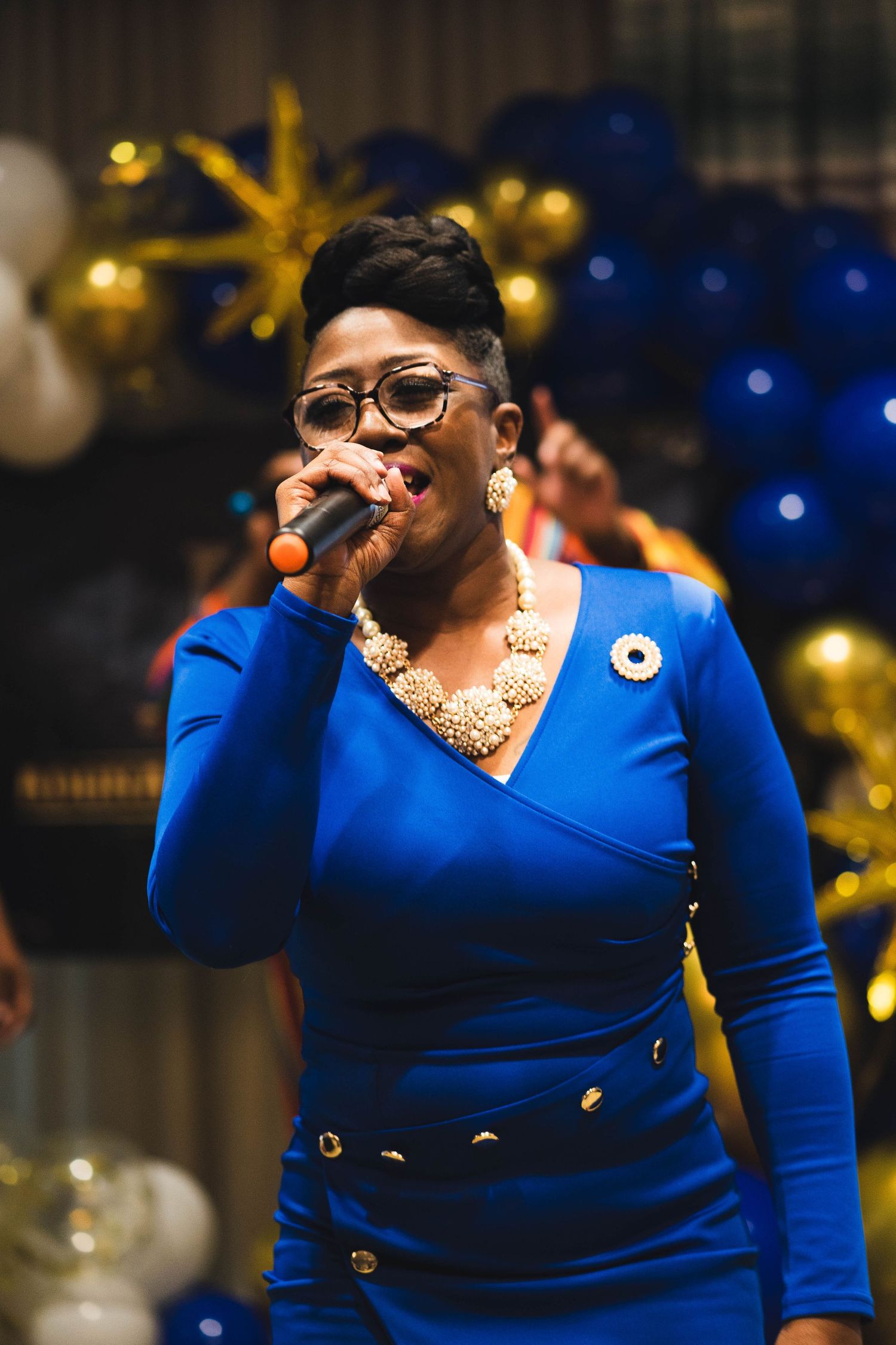 Performer in a blue dress singing on stage with festive gold decorations in the background.