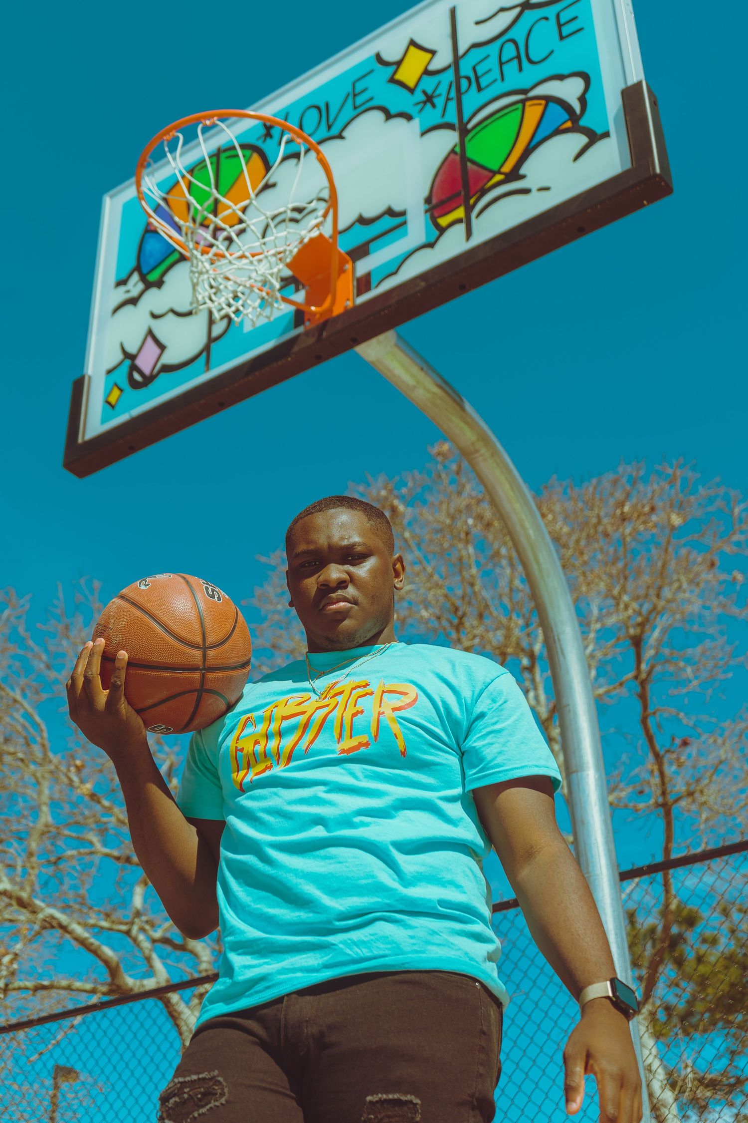Basketball player holding a ball under a colorful graffiti backboard against bright blue sky.