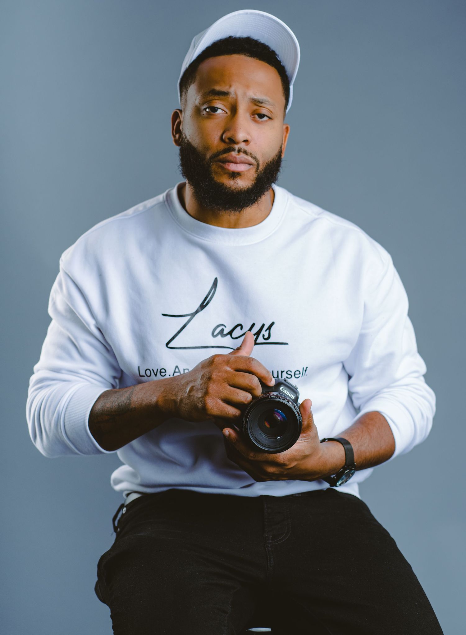 Photographer wearing a white sweatshirt and baseball cap holding a camera against a gray backdrop.