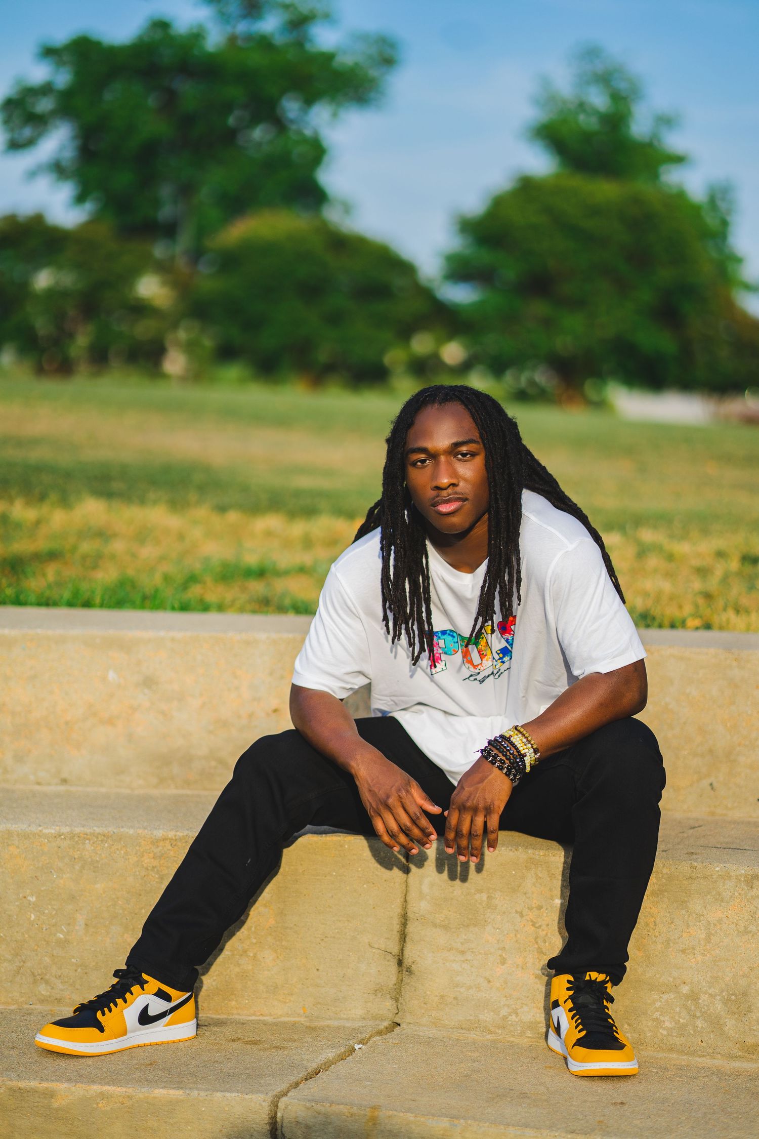 Casual portrait of someone in white t-shirt and black pants sitting outdoors wearing yellow Nike sneakers.