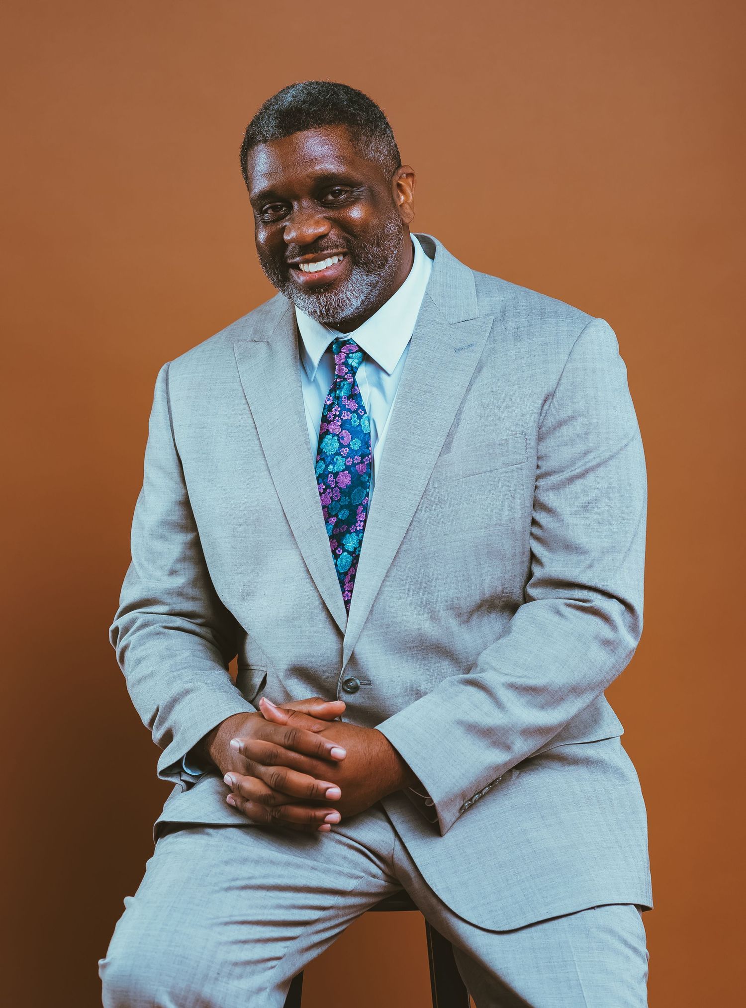 Professional portrait of a person in a light gray suit and floral tie against a brown background.