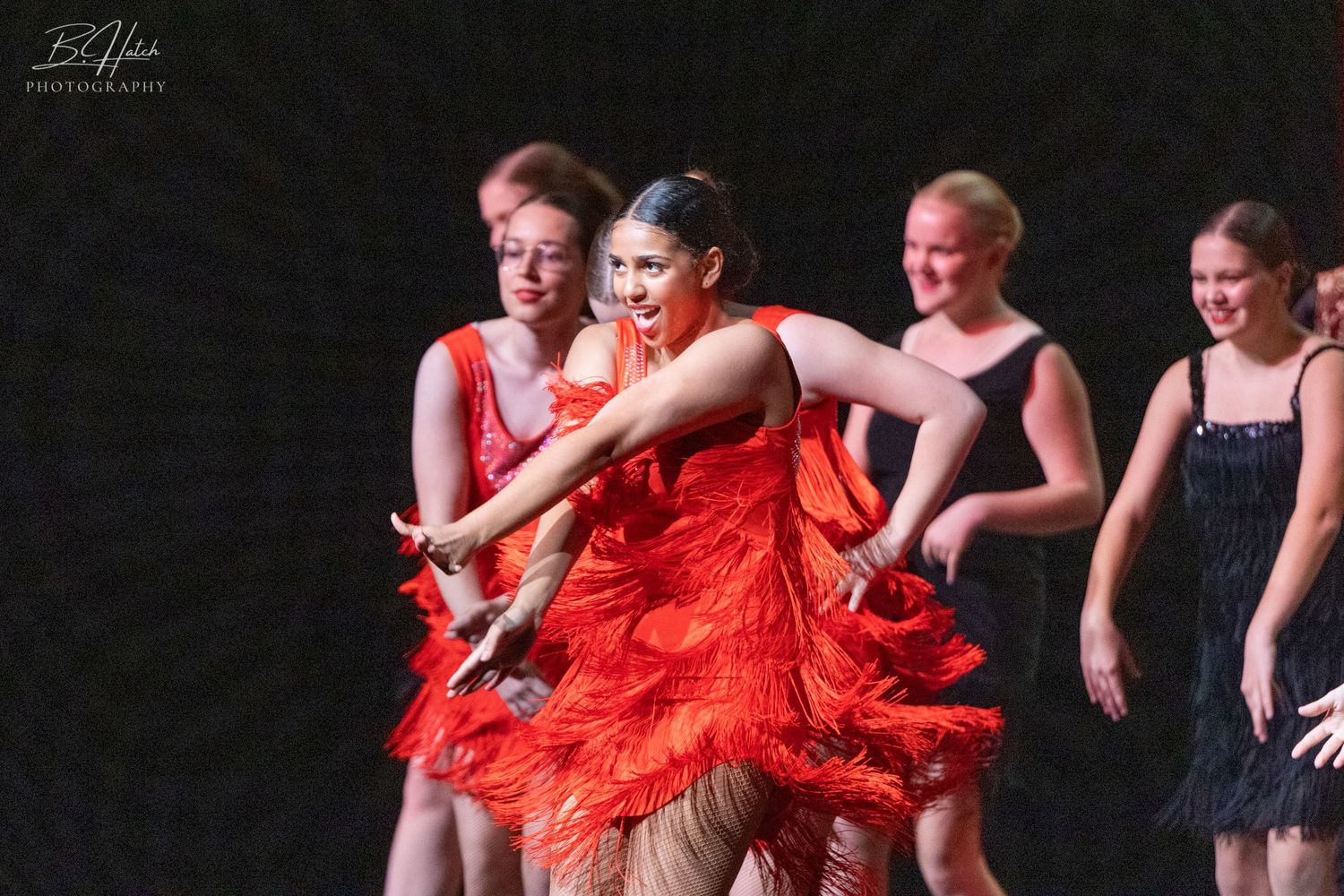 Dancers in red fringe costumes perform synchronized dance moves on dark stage.