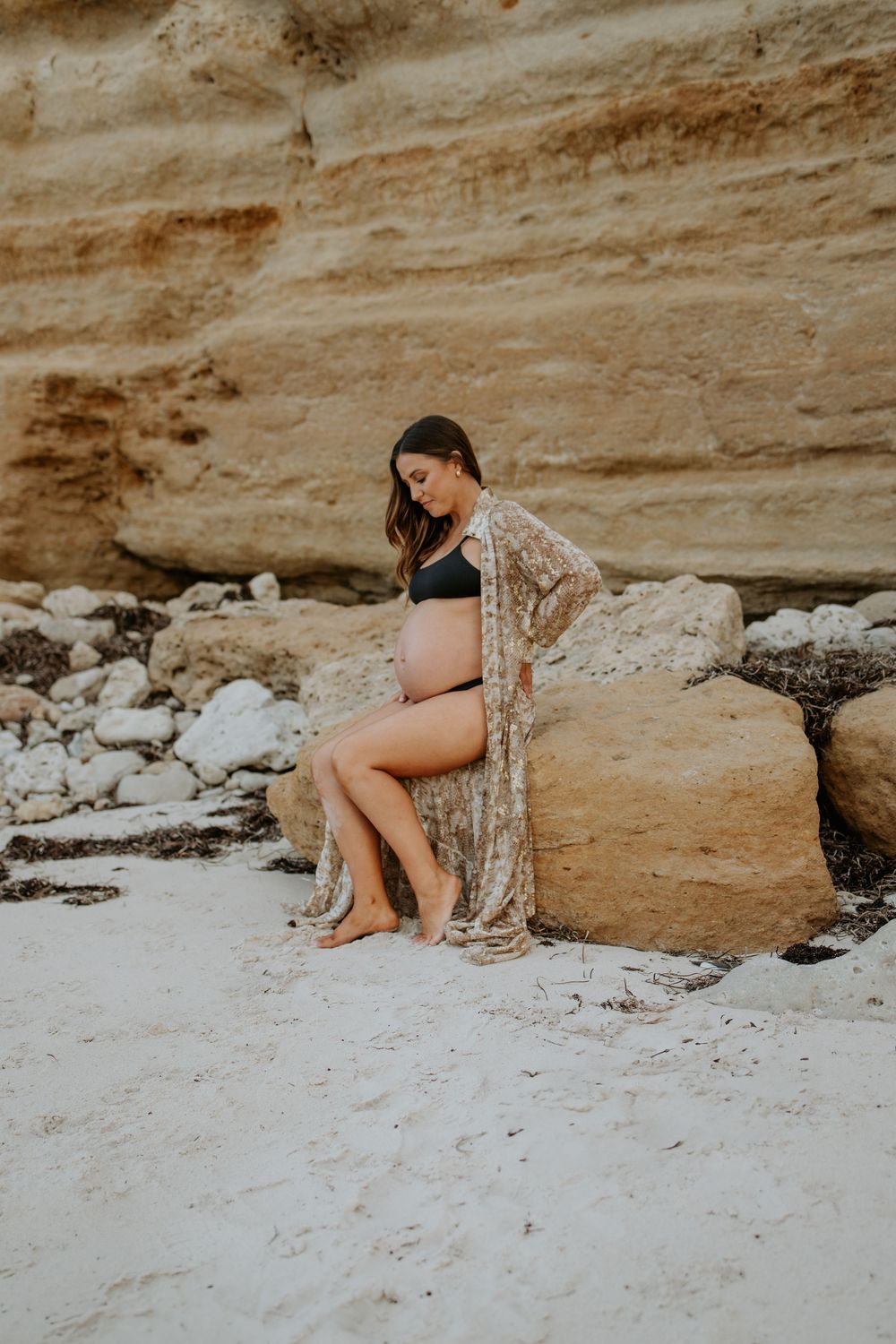 Expecting mum sitting against the cliff face during a natural sunset pregnancy photography session at Port Willunga, South Australia