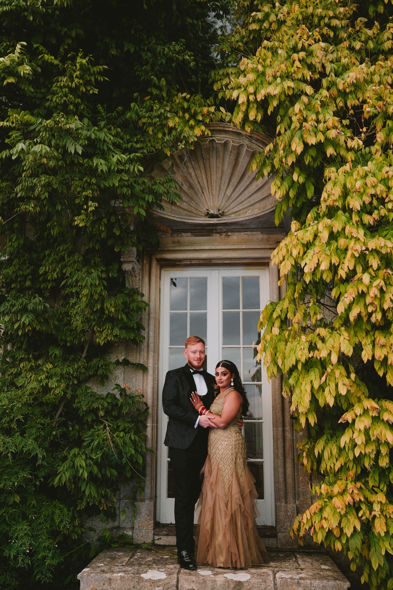 Couple poses in formal attire against an ivy-covered doorway with vintage architectural details.
