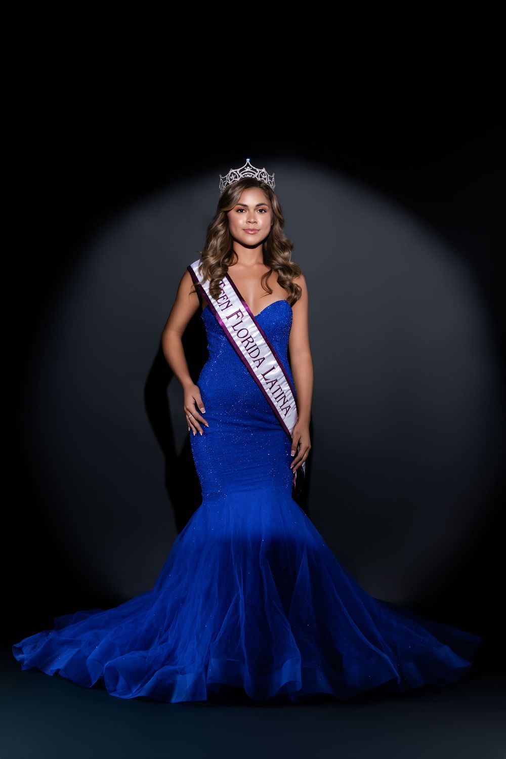 Beauty pageant contestant poses in a flowing royal blue gown with sash and crown.