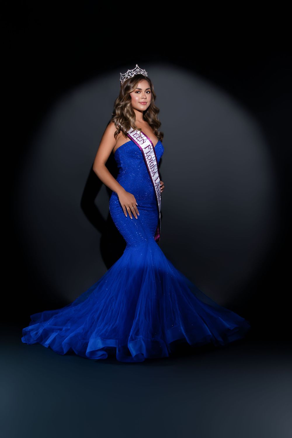 Dramatic photoshoot of pageant winner in sparkling blue ball gown against dark backdrop.