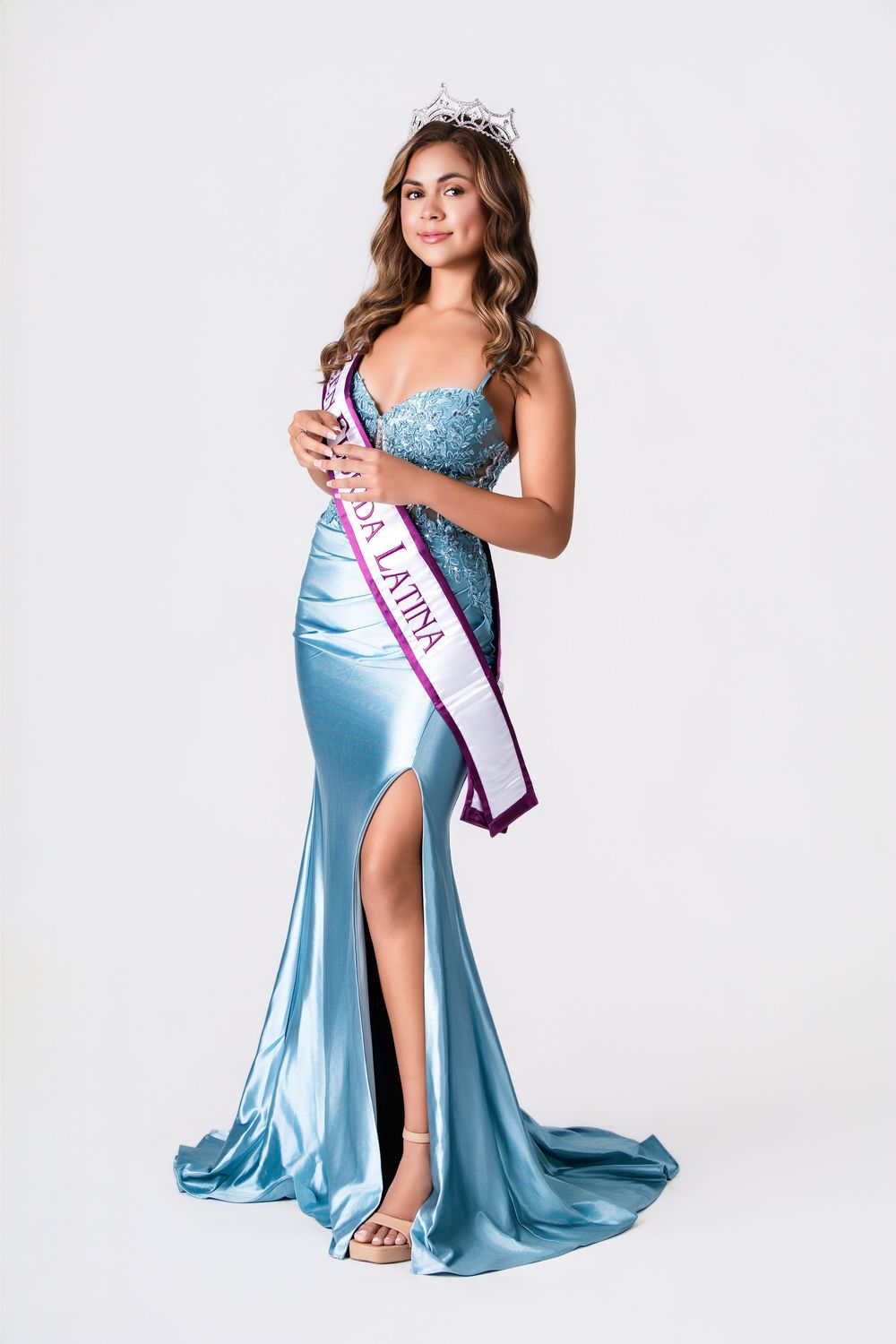 Beauty pageant contestant wearing a light blue satin gown and crown poses elegantly against white backdrop.