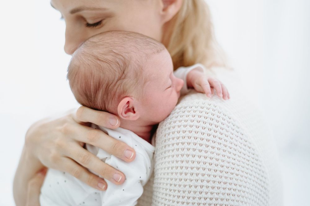 Mother gently holds sleeping newborn baby against her shoulder in soft white nursery setting.