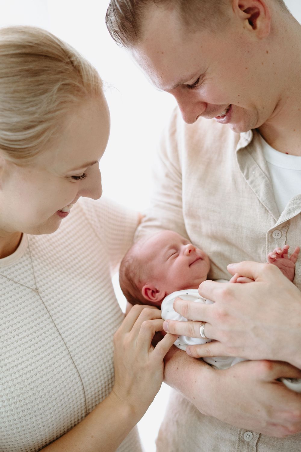 Happy parents smiling at their newborn baby, mother in pink top and father in beige shirt holding infant together.